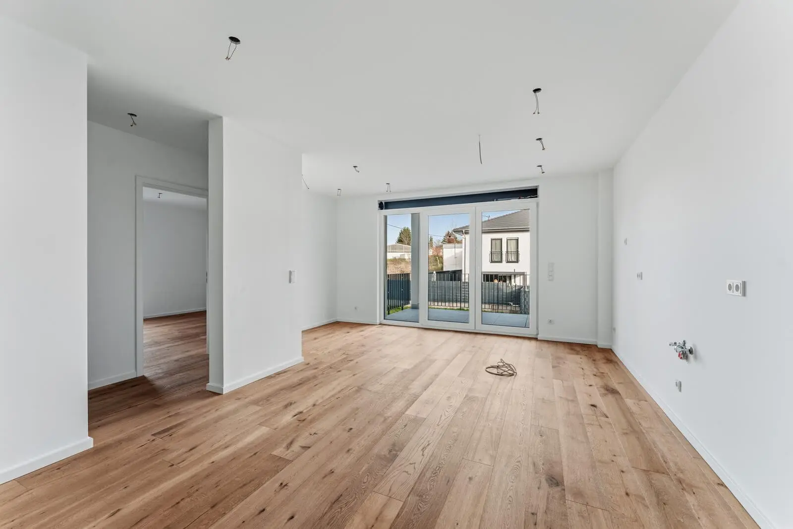 Leeres, unmöbliertes Zimmer mit weißen Wänden, Hartholzböden und großen Glasschiebetüren, die in einen umzäunten Hof führen; freiliegende Leuchten an der Decke.