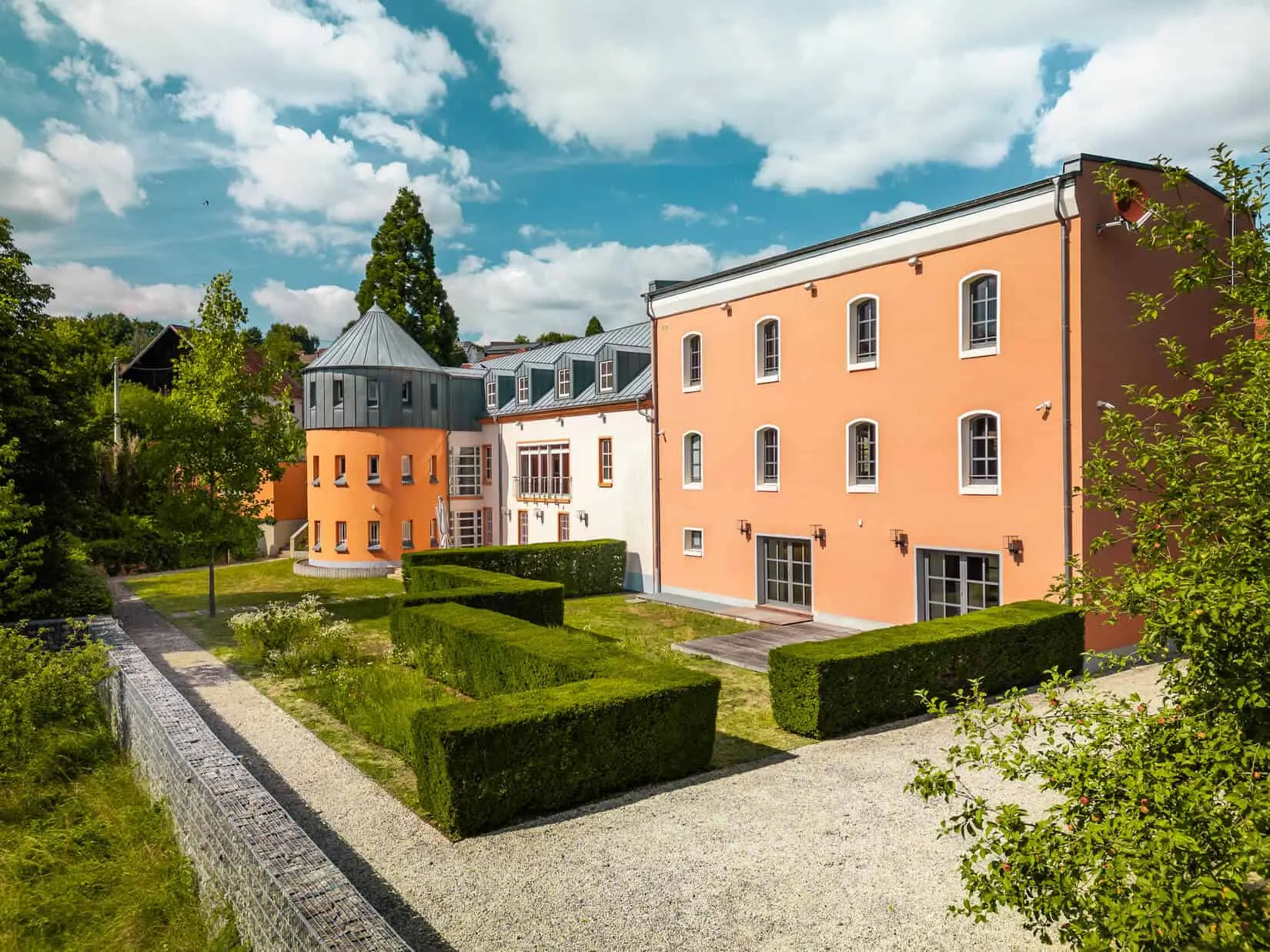 Modernes, orange- und cremefarbenes Gebäude mit großen Fenstern und einem runden Turm, hinter ordentlich geschnittenen Hecken und Kieswegen, unter einem teilweise bewölkten Himmel.