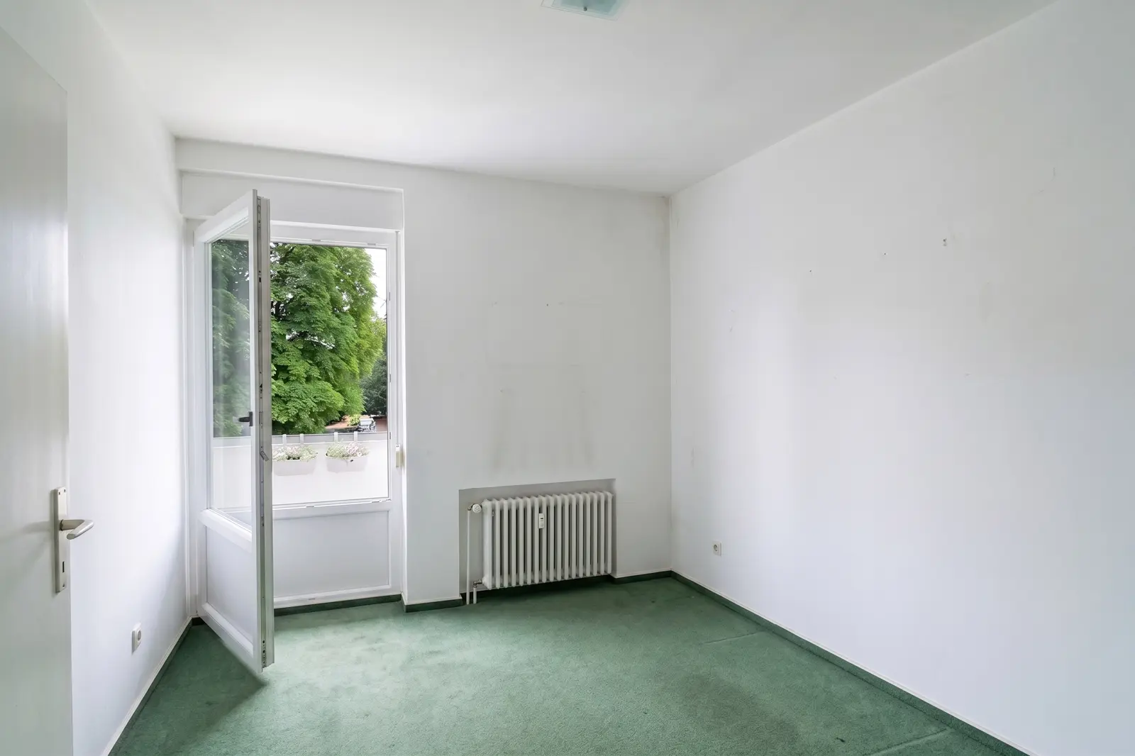 Ein leerer, weißer Raum mit grünem Teppich, einem Heizkörper und einem offenen Fenster, durch das man Bäume und einen Balkon sieht.