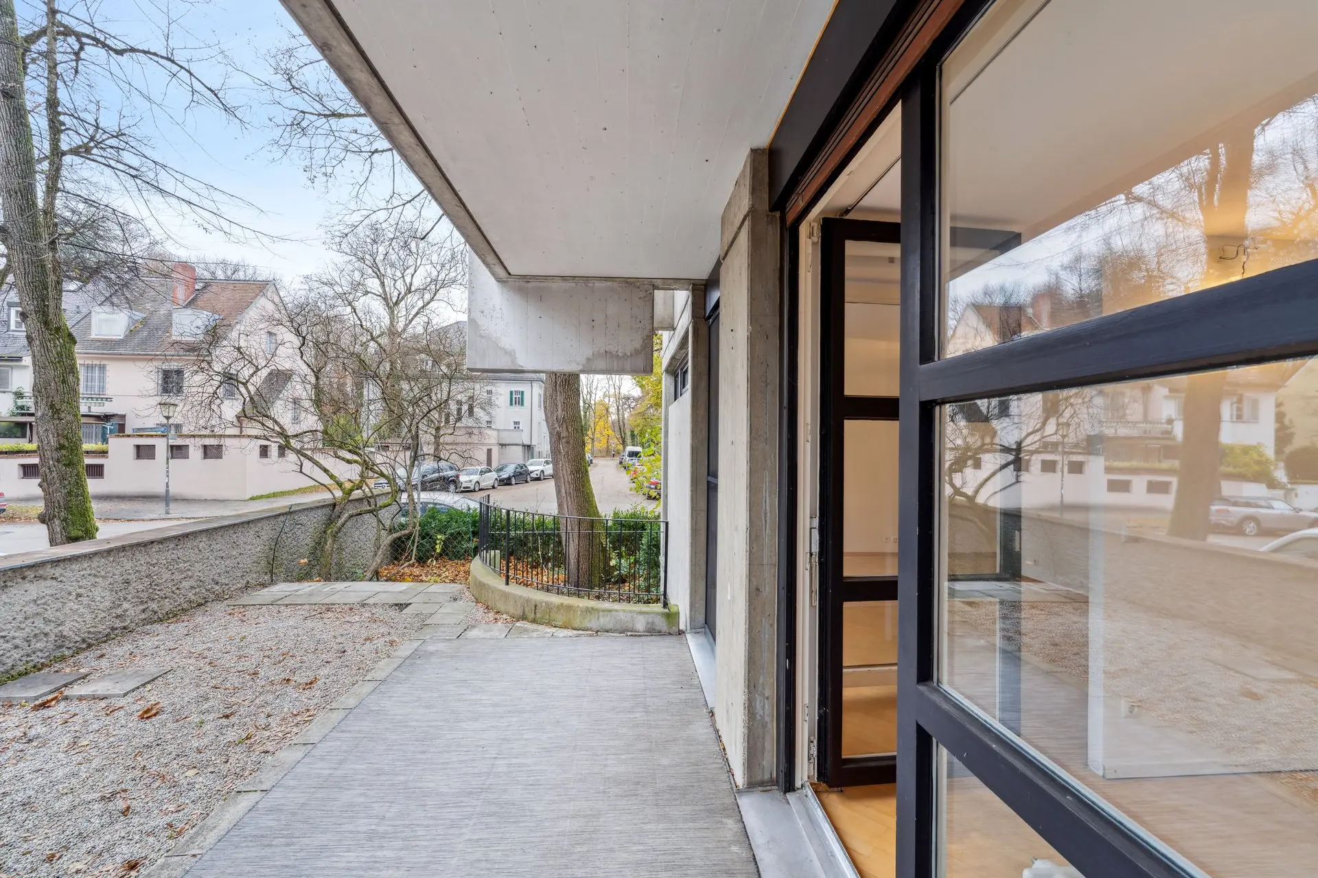 Ein moderner, offener Eingang mit Glastüren führt zu einer Betonterrasse und erschließt den Charme dieser 2-Zimmer-Wohnung in Bogenhausen. Blattlose Bäume und Wohnhäuser bilden eine ruhige Kulisse.