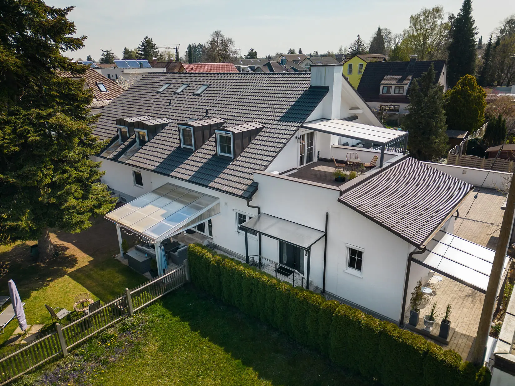 Luftaufnahme eines modernen zweistöckigen weißen Hauses mit mehreren überdachten Terrassen, einem umzäunten Garten und Nachbarhäusern im Hintergrund.