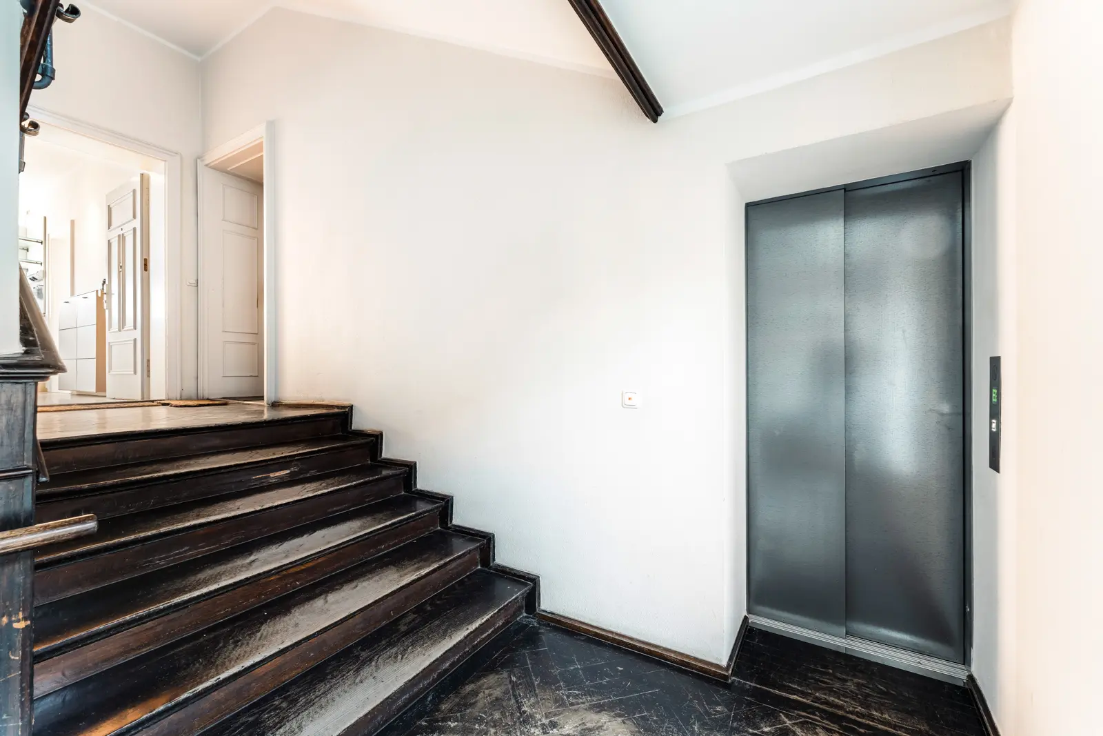 Ein kleiner Aufzug mit geschlossenen Metalltüren neben einer dunklen Holztreppe in einem hell beleuchteten Flur mit weißen Wänden.
