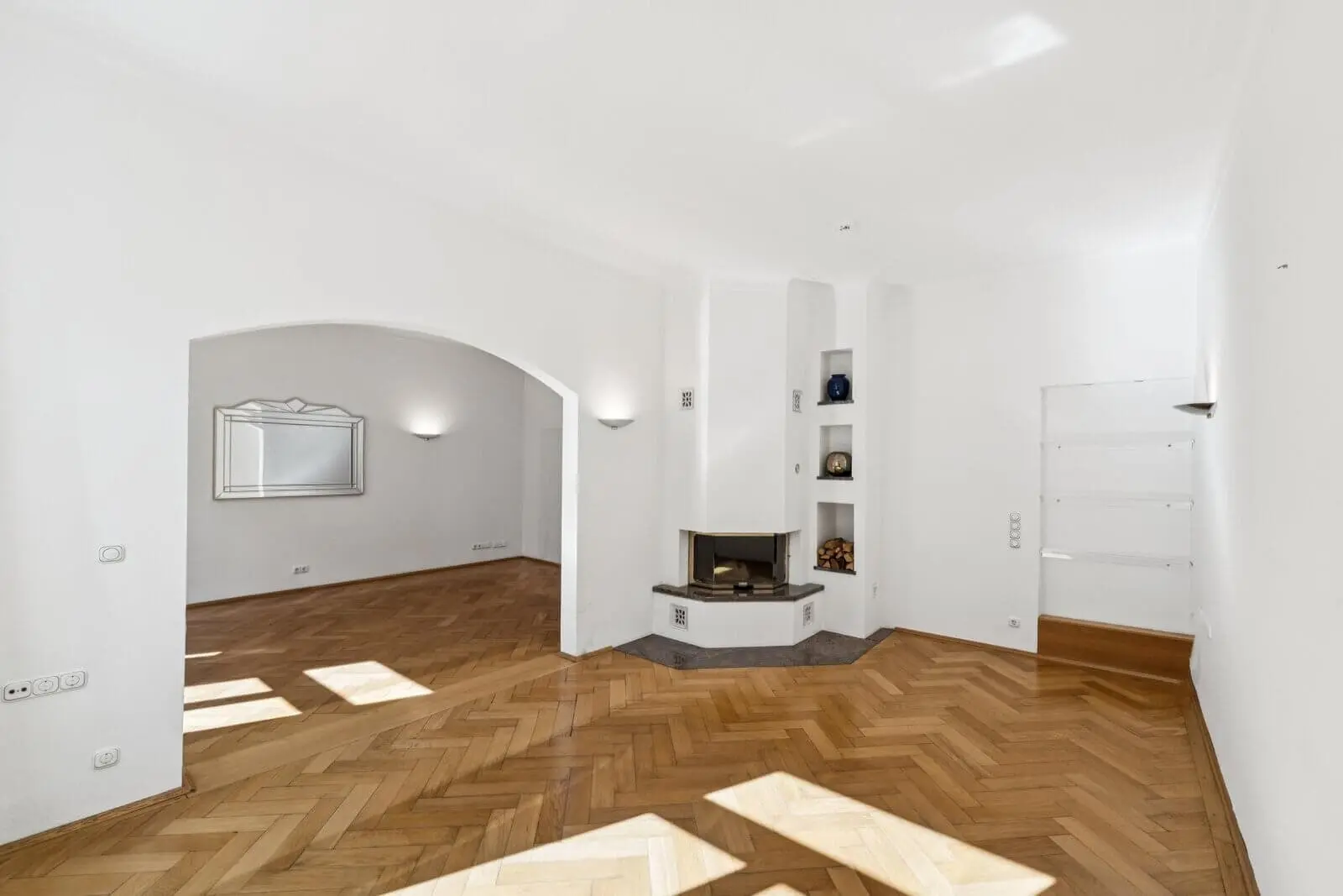 Heller, leerer Raum mit Fischgrätparkett, Eckkamin, Einbauregalen, gewölbter Tür und einem großen Wandspiegel.