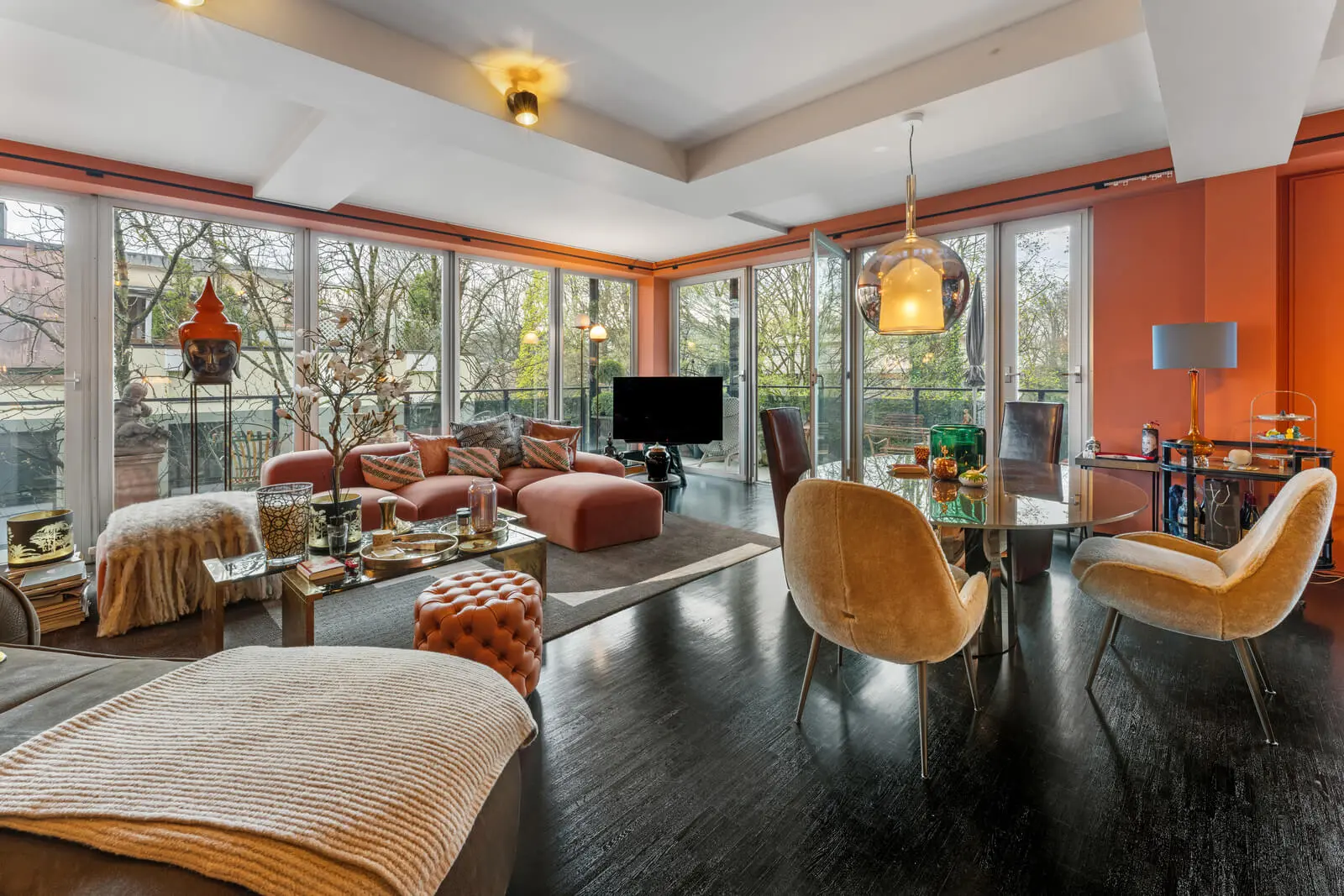 Ein modernes Wohnzimmer mit großen Fenstern, orangefarbenen Wänden, dunklem Holzboden, zeitgenössischen Möbeln darunter einem Fernseher, Sofas, Stühlen und verschiedenen Dekorationsgegenständen.