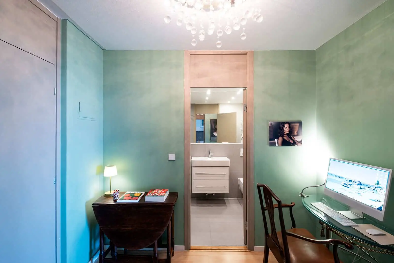 Kleines Heimbüro mit grünen Wänden, einem Glastisch mit Computer, einem Holzstuhl, einem Beistelltisch mit Büchern und Lampe und einem Blick durch eine offene Tür in ein modernes Badezimmer.