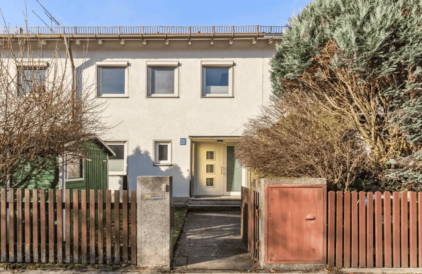 Ein zweistöckiges, weißes Wohnhaus mit vier Fenstern, einer grünen Tür und einem kleinen eingezäunten Vorgarten mit Bäumen und einem Schuppen.
