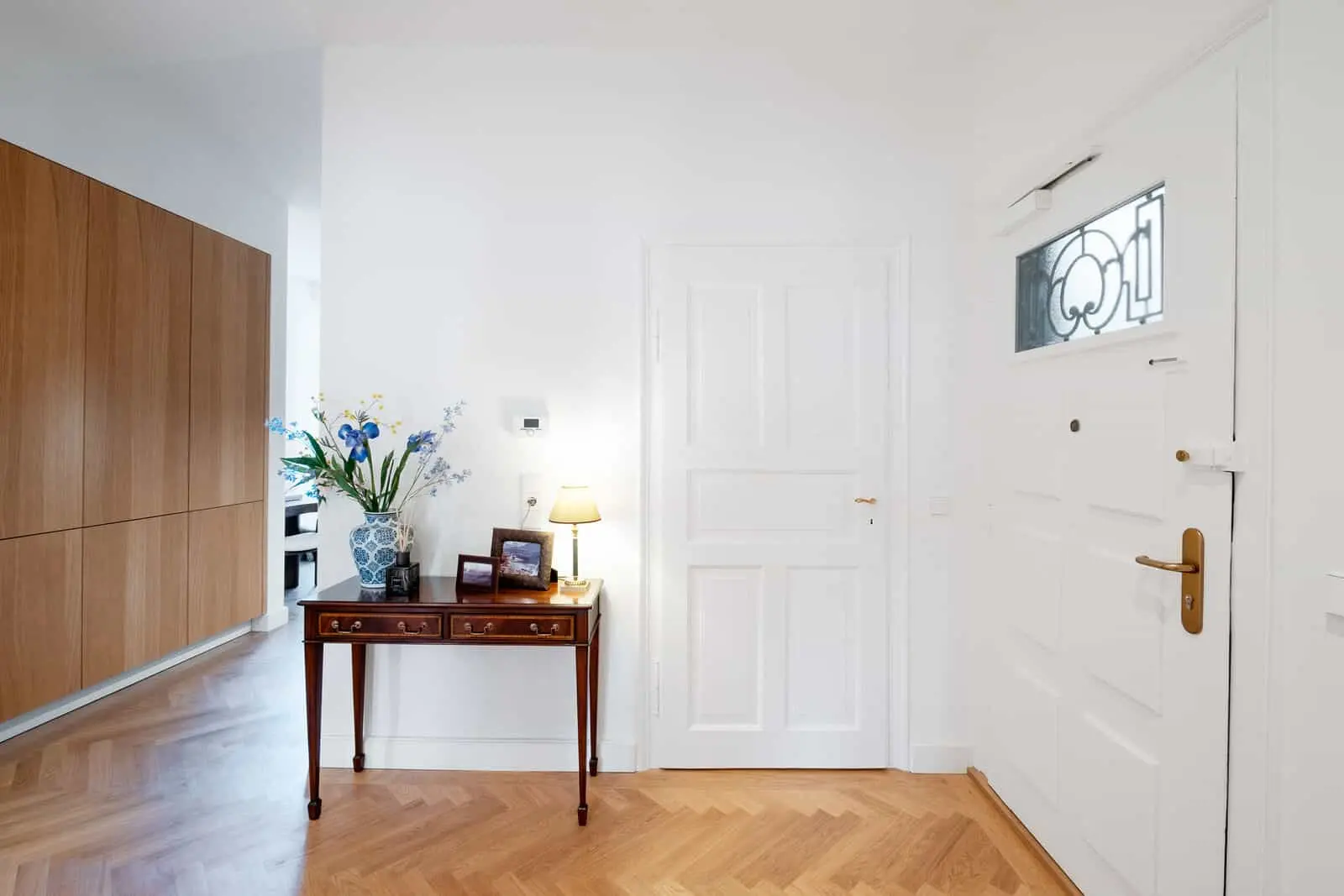 Ein weißer Flur mit einem Holzboden, einer Eingangstür, einem Beistelltisch mit einer Lampe, gerahmten Fotos und einer blauen Vase mit Blumen neben einem Holzeinbauschrank.