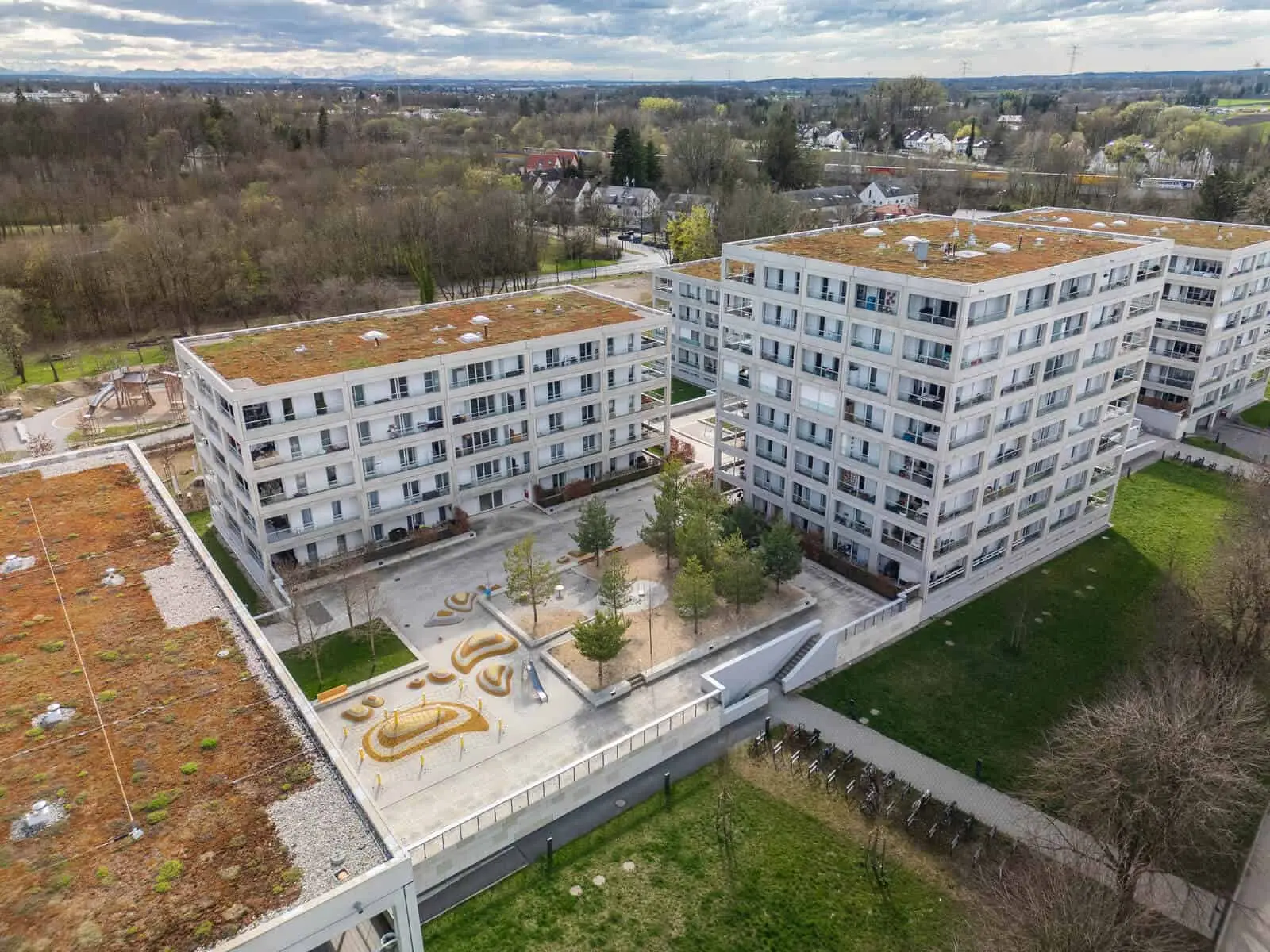 Luftaufnahme von modernen Wohngebäuden mit begrünten Dächern, einem zentralen Innenhof mit Bäumen und einem Spielplatz, umgeben von Grünanlagen und einem Park.