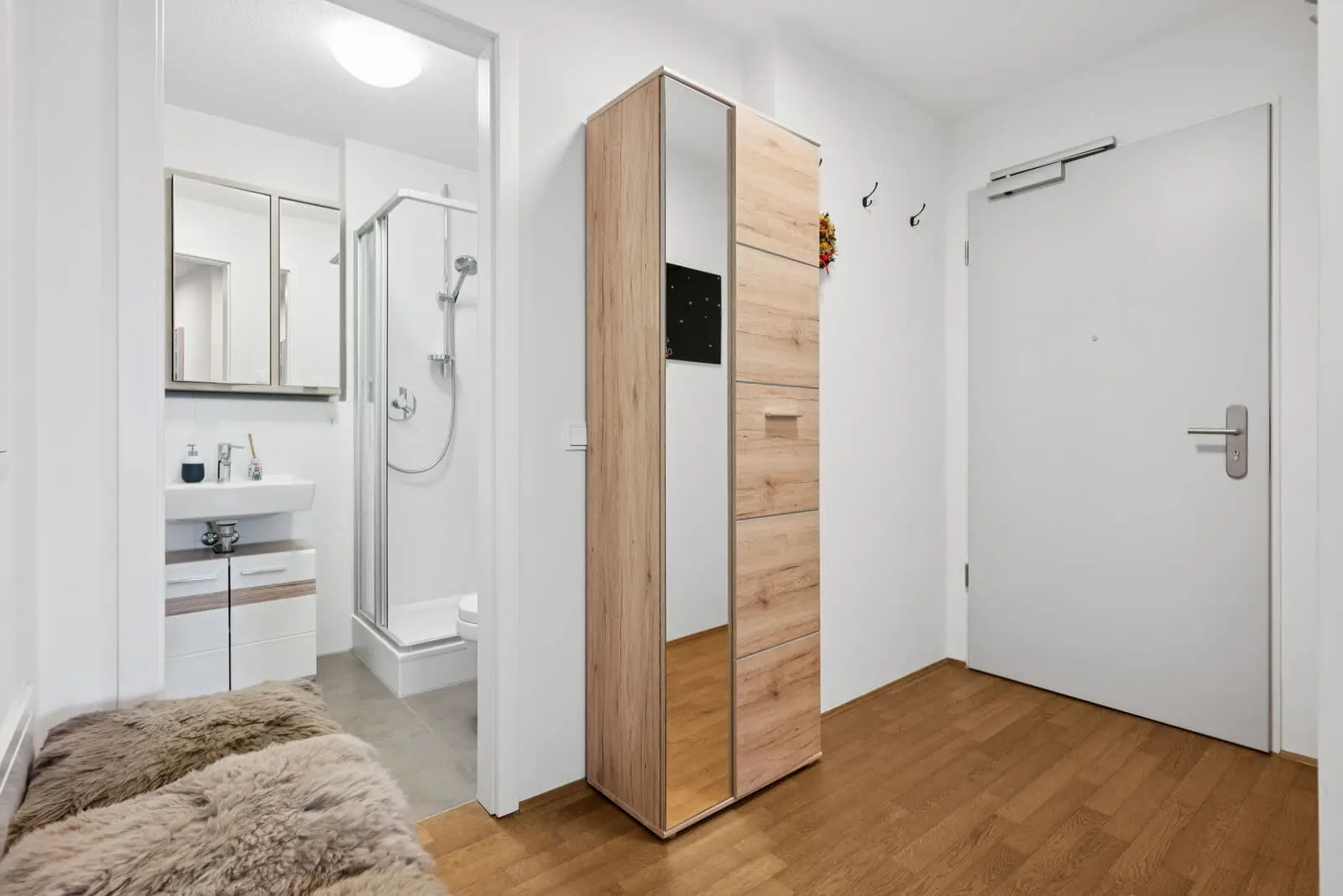 Moderner Eingangsbereich mit Holzboden, einem Kleiderschrank mit Spiegel, Wandhaken und einer Tür. Das Badezimmer mit weißen Armaturen und Glasdusche ist durch eine offene Tür sichtbar.
