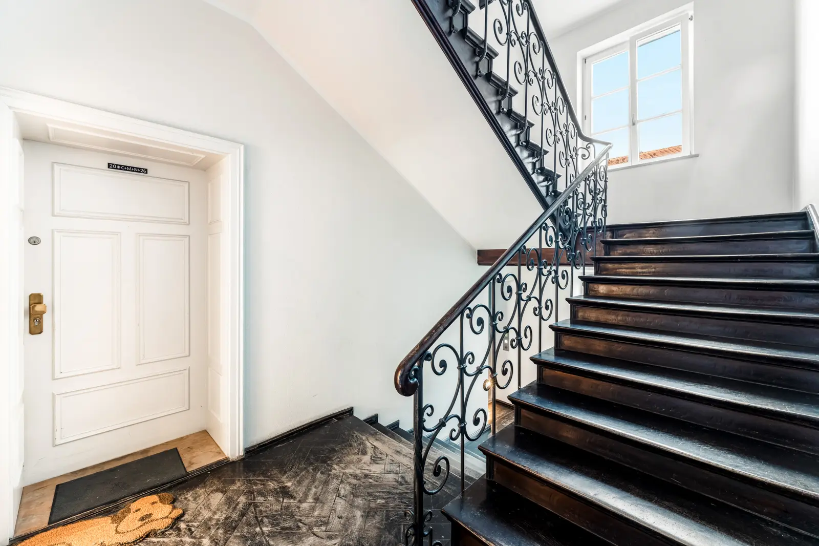 Eine schwarze Holztreppe mit verziertem Geländer neben einer weißen Tür in einem hellen Flur mit weißen Wänden und einem Fenster, durch das Tageslicht hereinfällt.