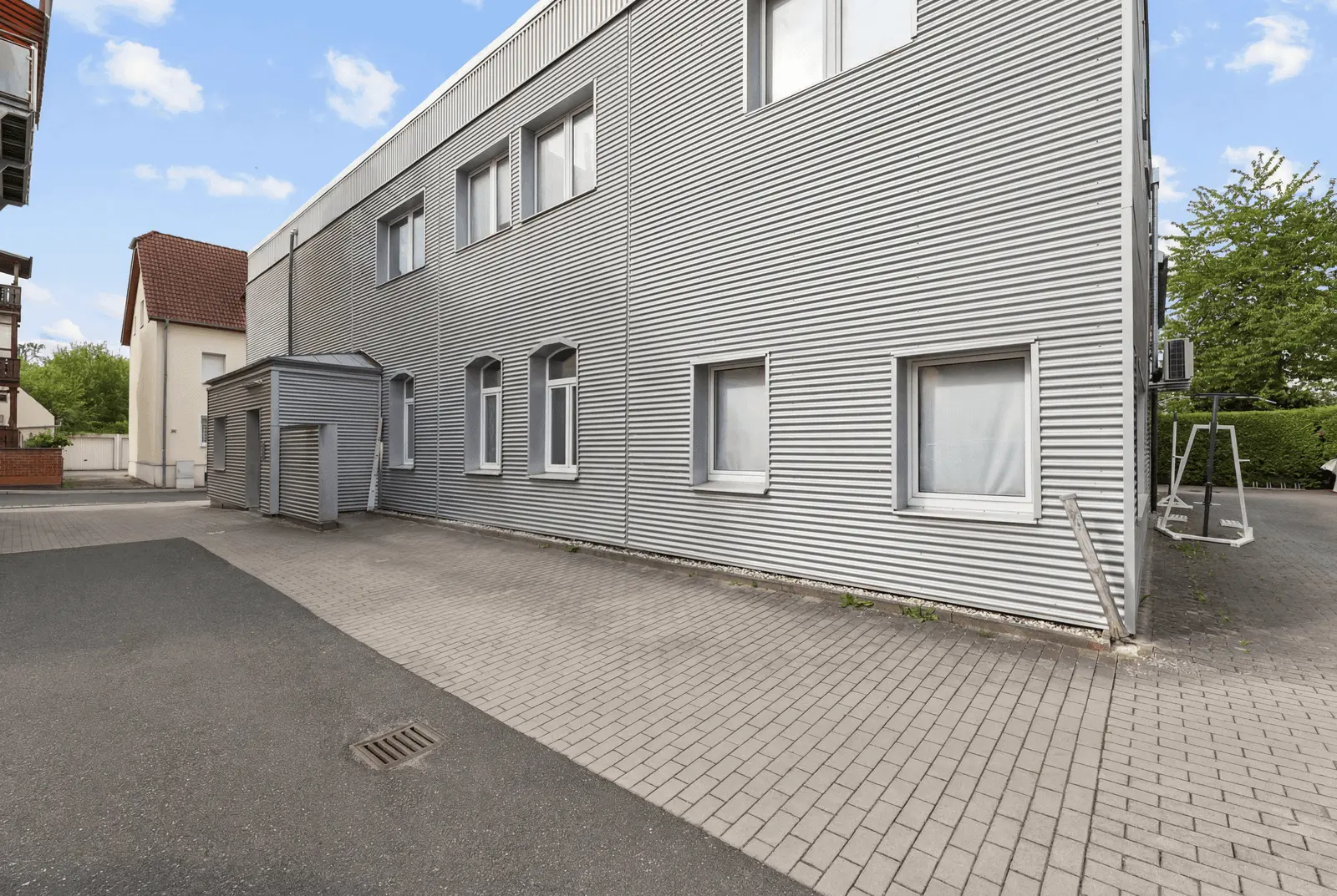 Ein modernes Industriegebäude mit Wellblechverkleidung, mehreren Fenstern und einem kleinen Anbau neben einer gepflasterten Zufahrt und grünen Bäumen im Hintergrund.