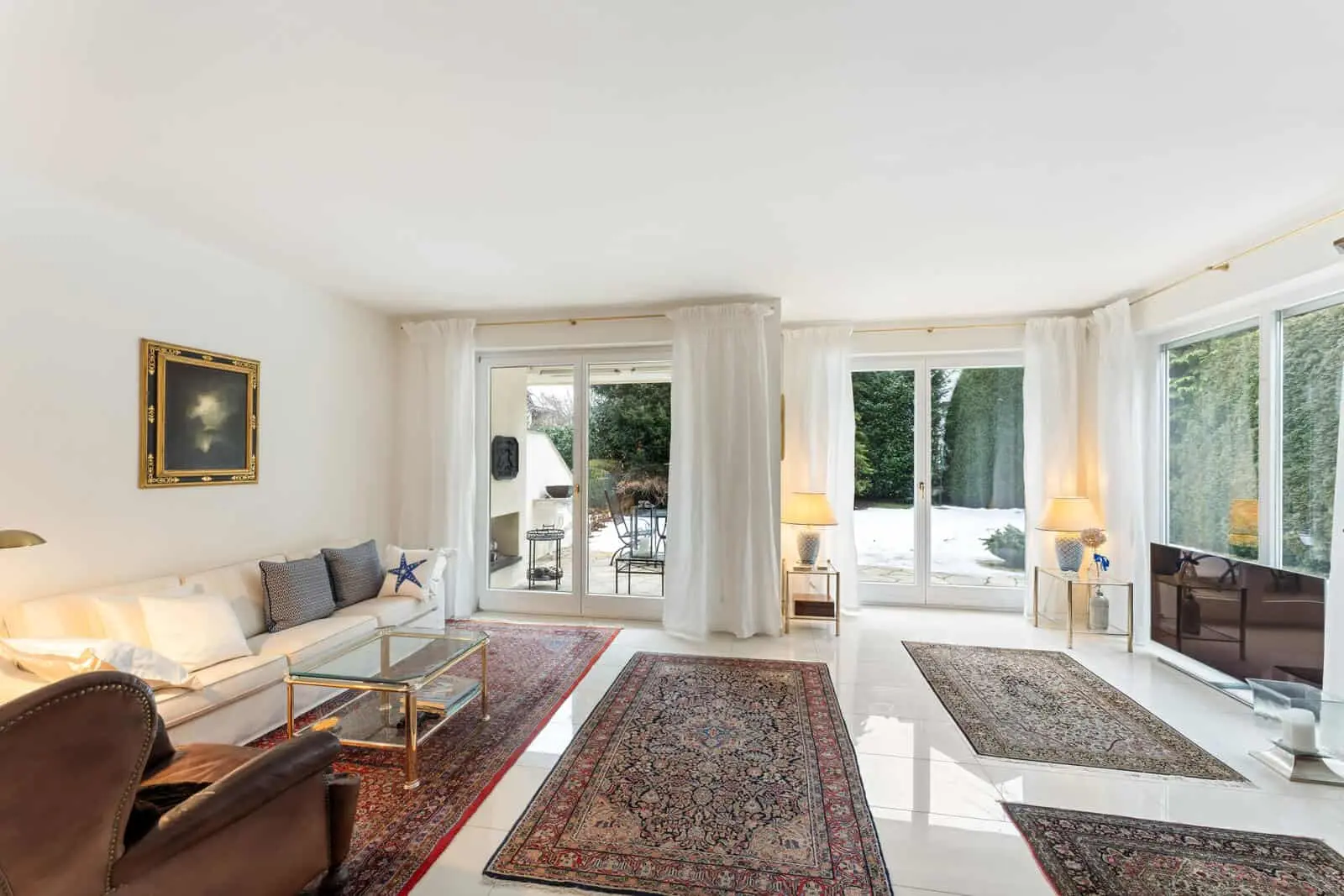 Helles Wohnzimmer mit großen Fenstern, Glastüren, weißen Wänden, gemusterten Teppichen, einem weißen Sofa, einem Ledersessel und Möbeln mit Goldakzenten. Draußen ist die schneebedeckte Terrasse zu sehen.