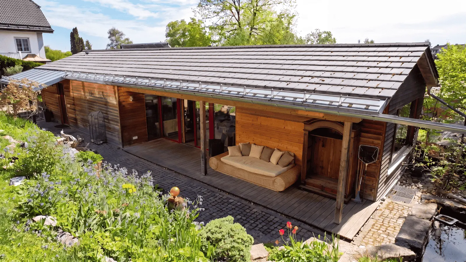 Einstöckiges Holzhaus mit einer überdachten Veranda, Sitzgelegenheiten im Freien und einem Garten mit verschiedenen Blumen und Pflanzen davor.