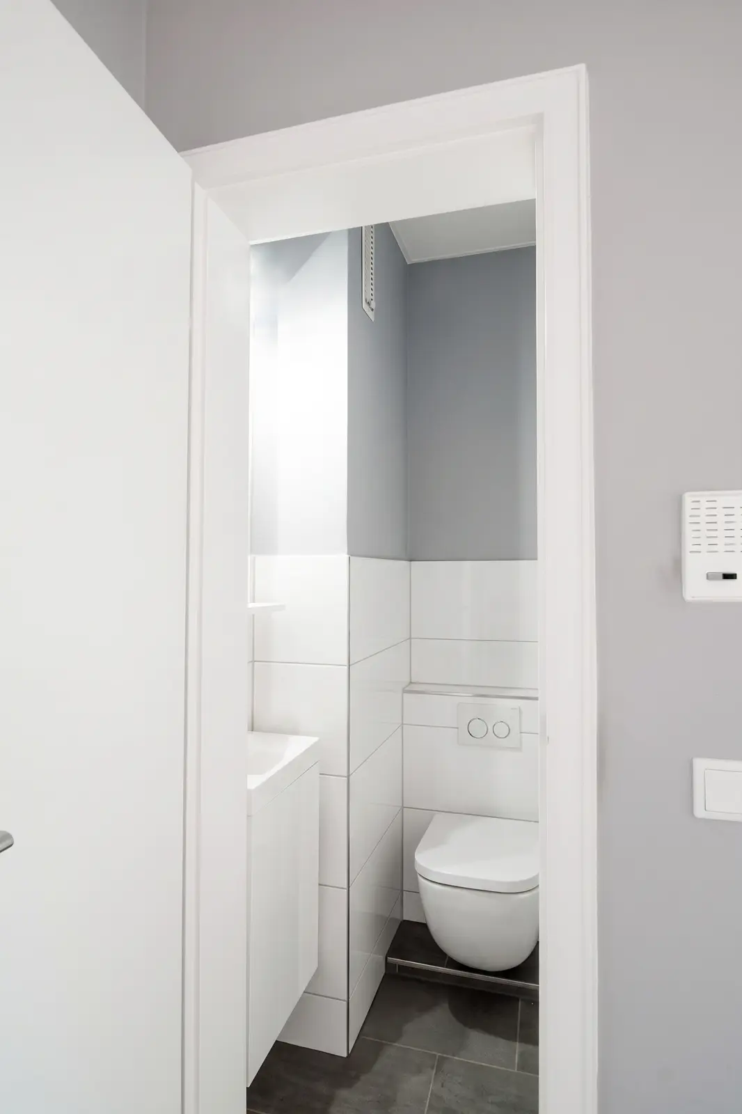Ein kleines, modernes Badezimmer mit einem wandhängenden WC, weißen Fliesen und grauen Wänden, gesehen durch eine teilweise geöffnete Tür.