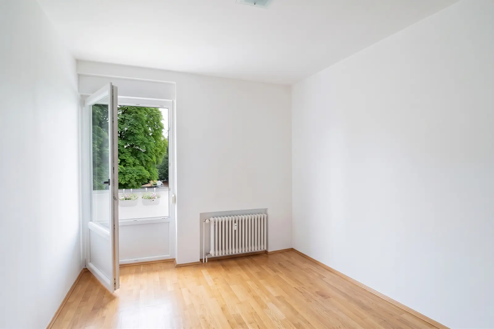 Ein leerer, weißer Raum mit Holzboden, offenem Fenster, Heizkörper und Blick auf grüne Bäume draußen.