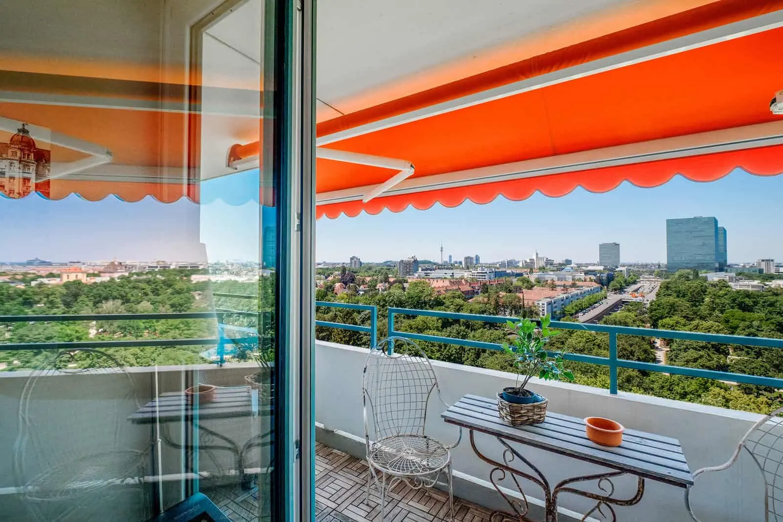 Ein Balkon mit einem kleinen Tisch, Stühlen und einer Topfpflanze bietet an einem sonnigen Tag einen Blick auf eine Stadtlandschaft mit Bäumen und Gebäuden unter einer orangefarbenen Markise.