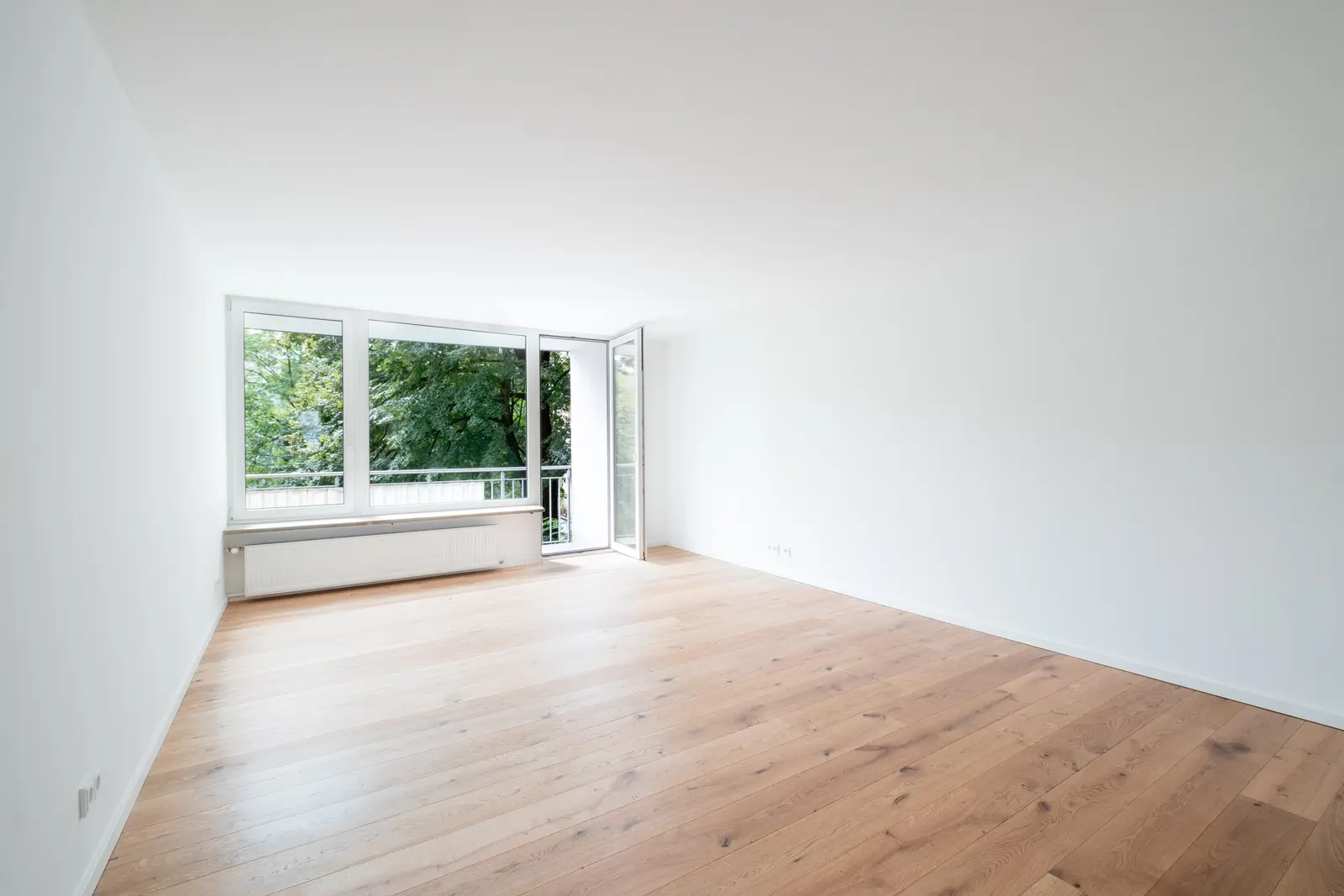 Ein leerer, heller Raum mit hellem Holzboden, weißen Wänden und großen Fenstern mit Blick auf grüne Bäume. Ein Fenster ist geöffnet, sodass Tageslicht in den Raum fällt.