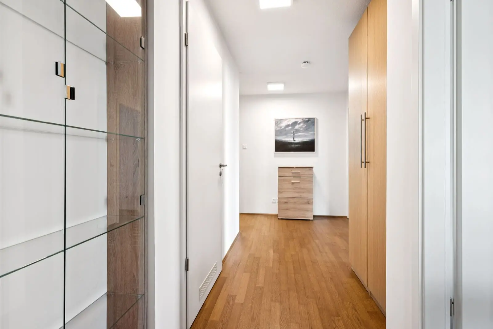 Ein moderner Flur mit Holzboden, hellen Wänden, Einbauschränken, einer Glasvitrine und einer kleinen Kommode mit einem gerahmten Bild darüber.