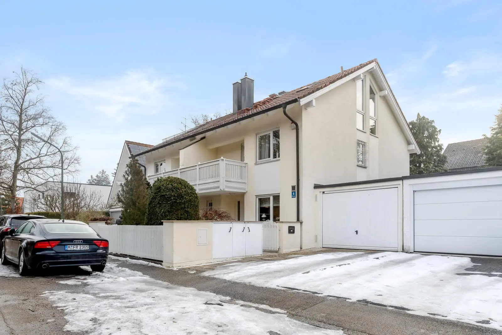 Zweistöckiges, cremefarbenes Haus mit Balkon, angebauter Garage und einer schneebedeckten Einfahrt. Ein schwarzes Auto ist auf der Straße vor dem Haus geparkt.