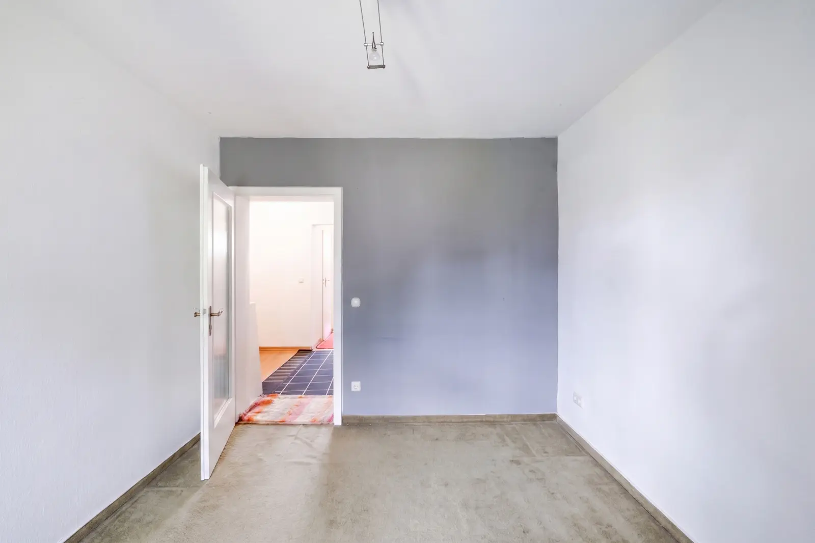 Ein leerer Raum mit grauem Teppichboden, einer grauen Akzentwand, einer offenen weißen Tür und Blick auf einen hell beleuchteten Flur mit Fliesenboden und einer weiteren Tür.