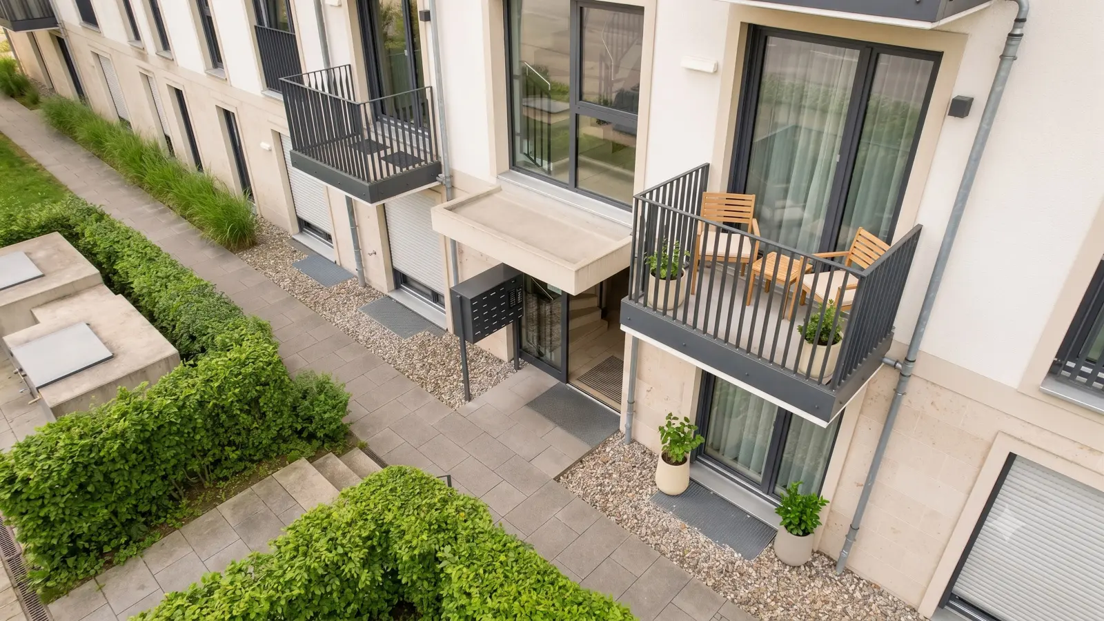 Fassade eines modernen Mehrfamilienhauses mit Balkonen, Topfpflanzen und einem gepflasterten Gehweg, der von grünen Hecken gesäumt ist.