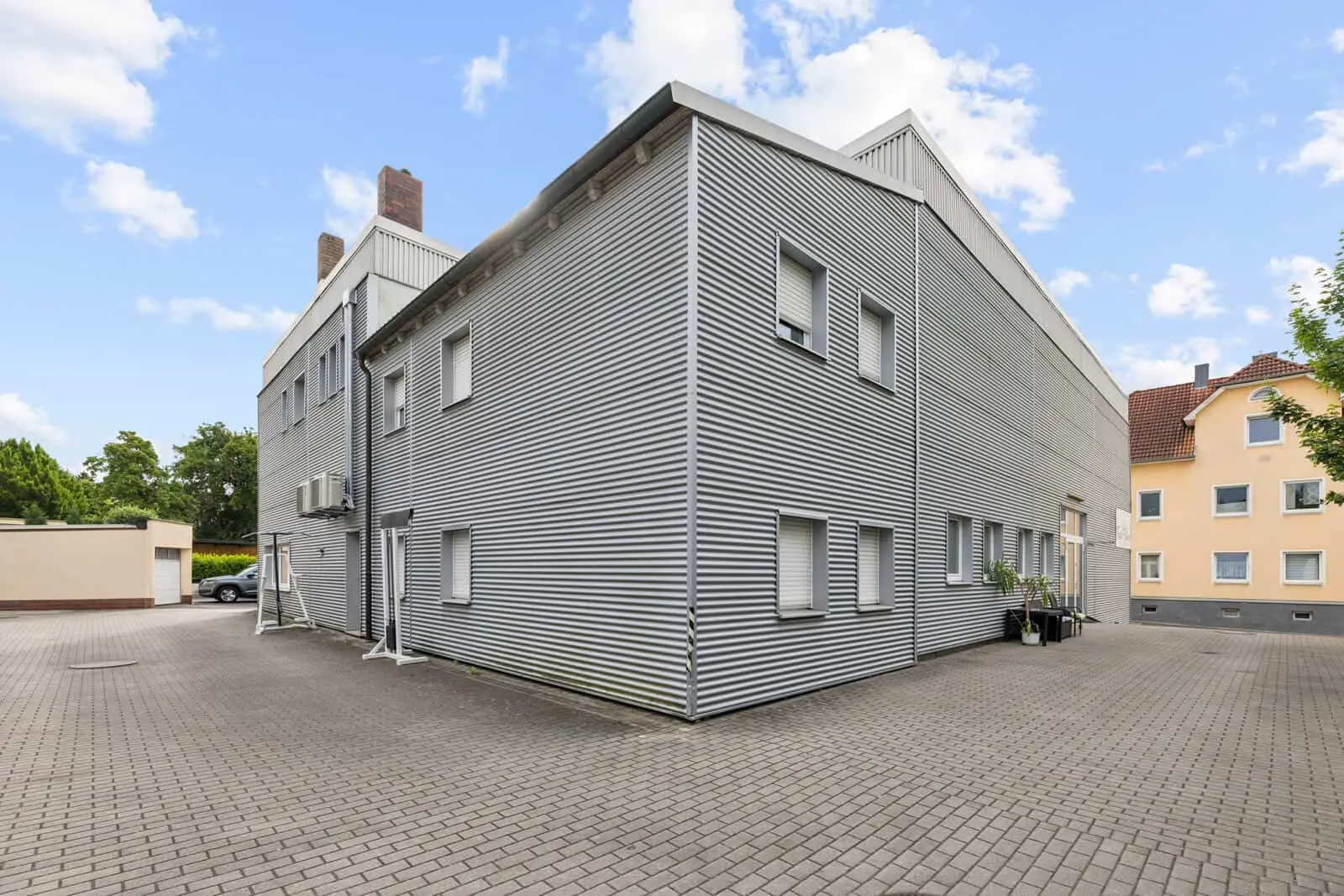 Ein modernes Industriegebäude mit Wellblechverkleidung, Flachdach und mehreren geschlossenen Fenstern, umgeben von einem gepflasterten Parkplatz unter einem teilweise bewölkten Himmel.