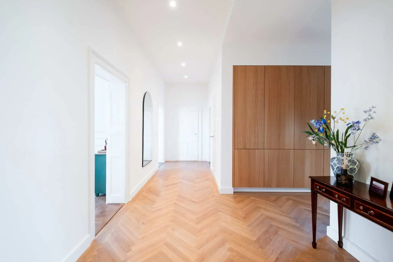 Heller Flur mit Holzfußboden im Fischgrätmuster, einem großen Spiegel an der linken Wand, einem Holzschrank auf der rechten Seite und einem Tisch mit einem Blumenarrangement. Mehrere Türen sind sichtbar.