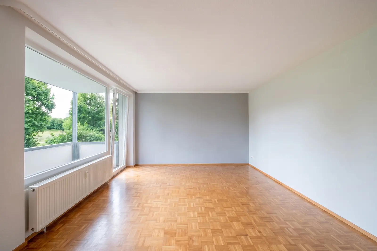 Ein leeres Wohnzimmer mit Parkettboden, einem großen Fenster, weißen Wänden, einer grauen Akzentwand und Blick ins Grüne.