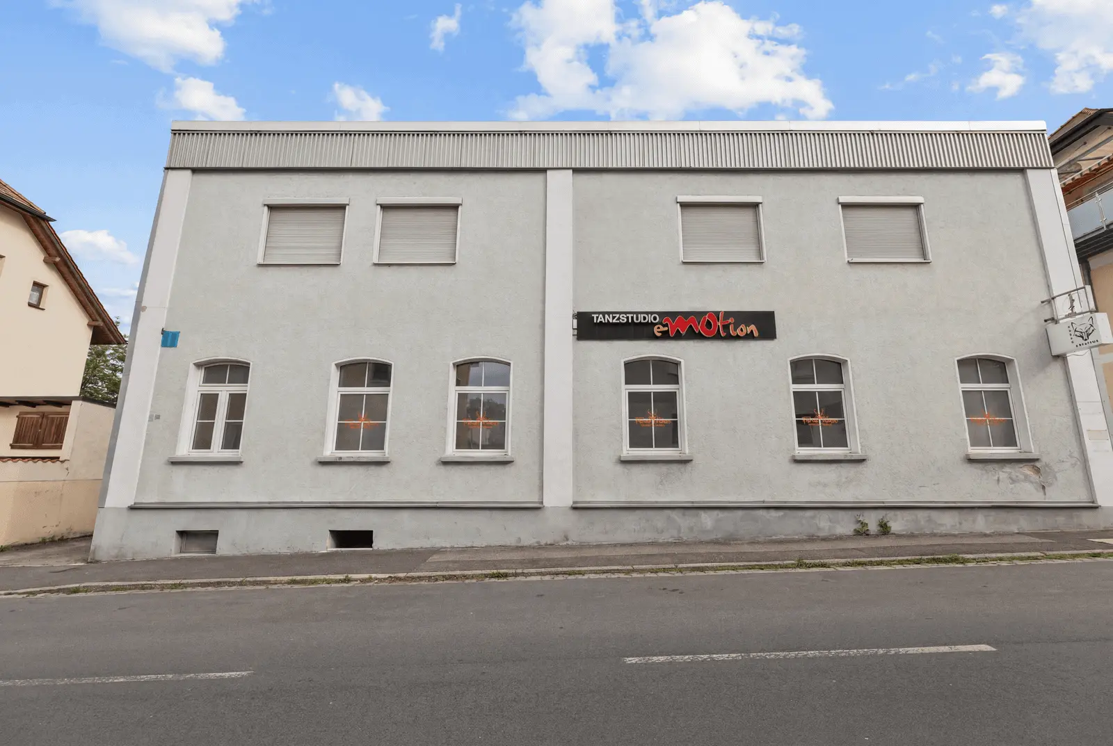 Ein hellgraues, zweistöckiges Gebäude mit sechs geschlossenen Fenstern im oberen Stockwerk, fünf Fenstern im unteren Stockwerk und einem Schild mit der Aufschrift "Tanzstudio eMotion" über der Tür.