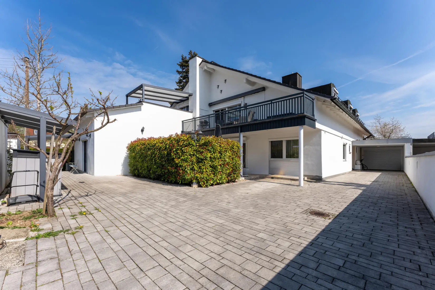 Modernes weißes zweistöckiges Haus mit großer gepflasterter Auffahrt, Balkon, Garage und geschnittenen Hecken unter einem klaren blauen Himmel.