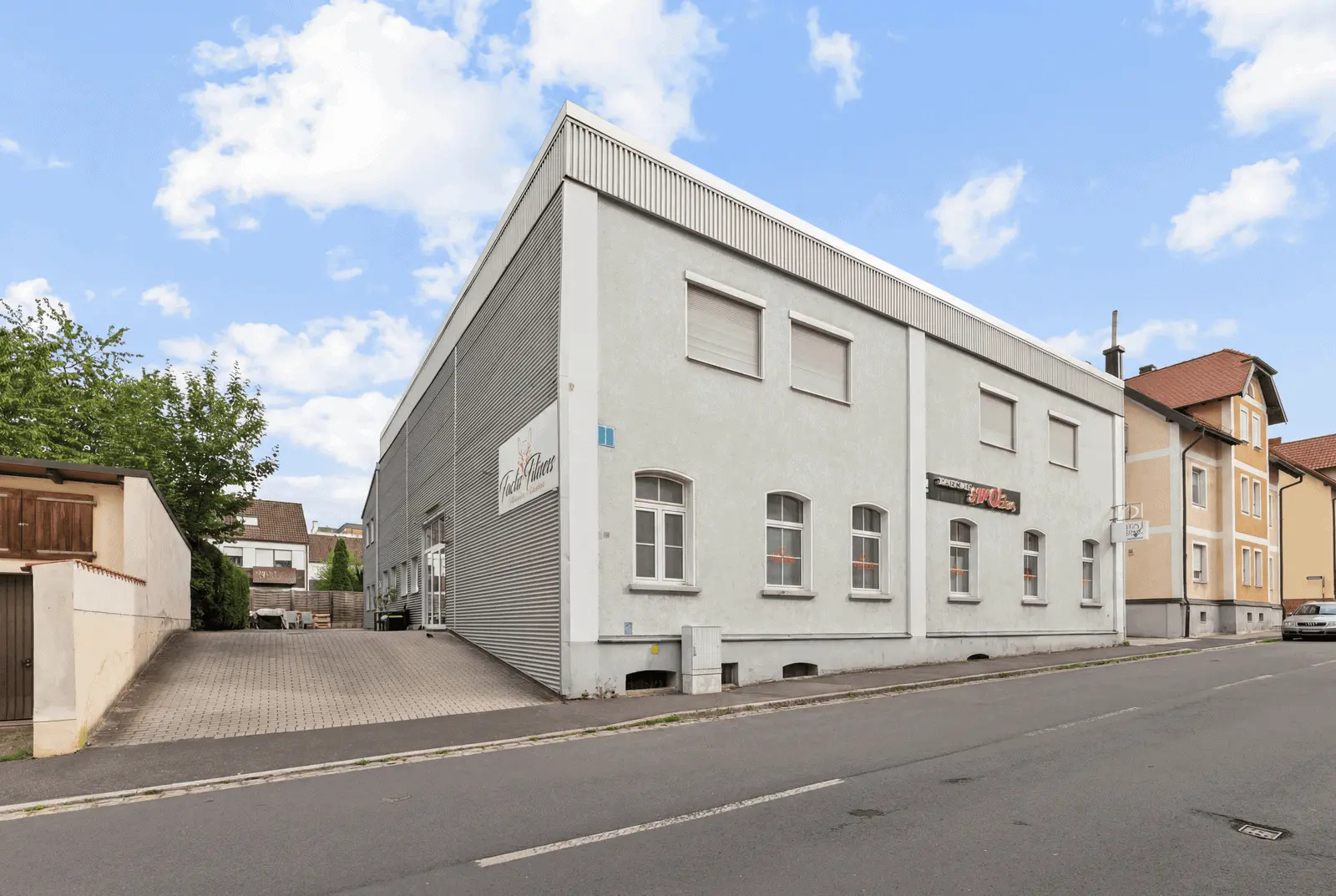 Ein großes, hellgraues Geschäftsgebäude mit mehreren Fenstern und einem Garagentor steht in einer ruhigen Straße neben Wohnhäusern unter einem teilweise bewölkten Himmel.