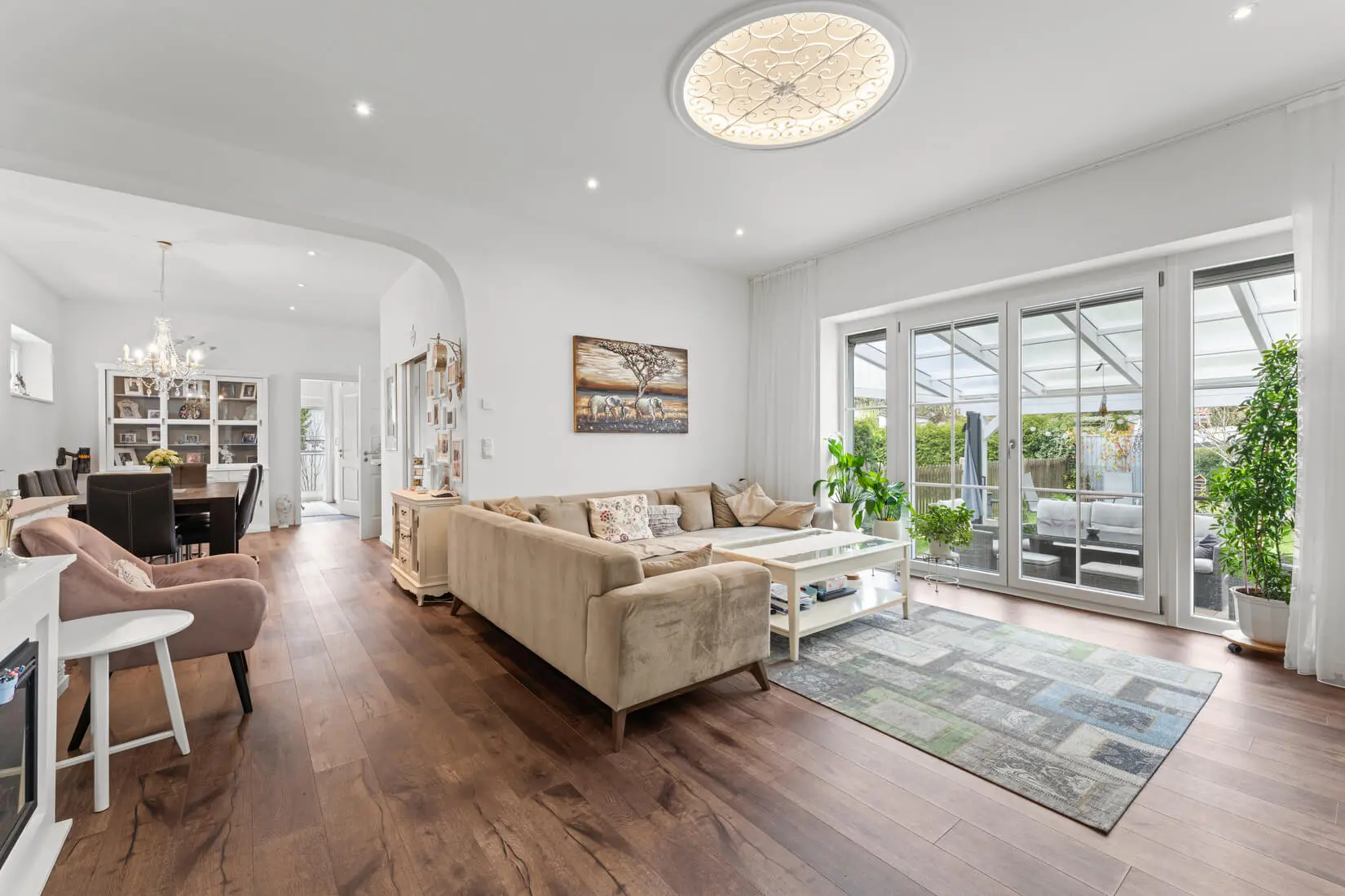 Modernes Wohnzimmer mit beigem Sofagarnitur, Holzböden und großen Fenstern, die zu einem hellen Wintergarten führen; im Hintergrund ist der Essbereich sichtbar.