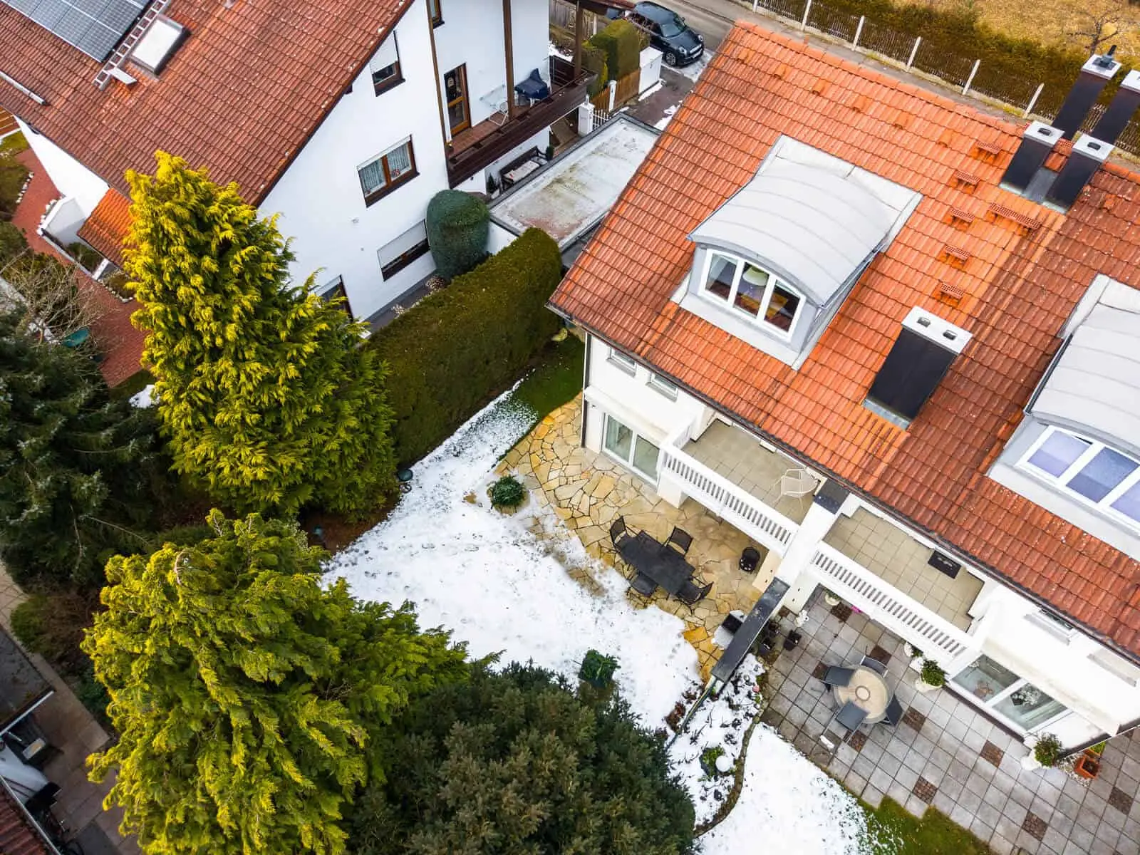 Luftaufnahme von zwei Häusern mit roten Ziegeldächern, einem teilweise schneebedeckten Hinterhof, Terrassenmöbeln und immergrünen Bäumen in der Umgebung.