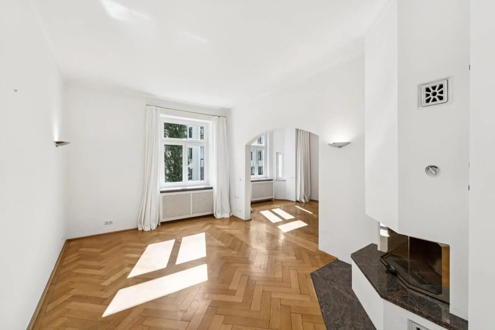 Heller, leerer Raum mit Parkettboden, weißen Wänden, großen Fenstern mit Vorhängen, einer gewölbten Tür und einem Eckkamin mit dunklem Steinsockel.