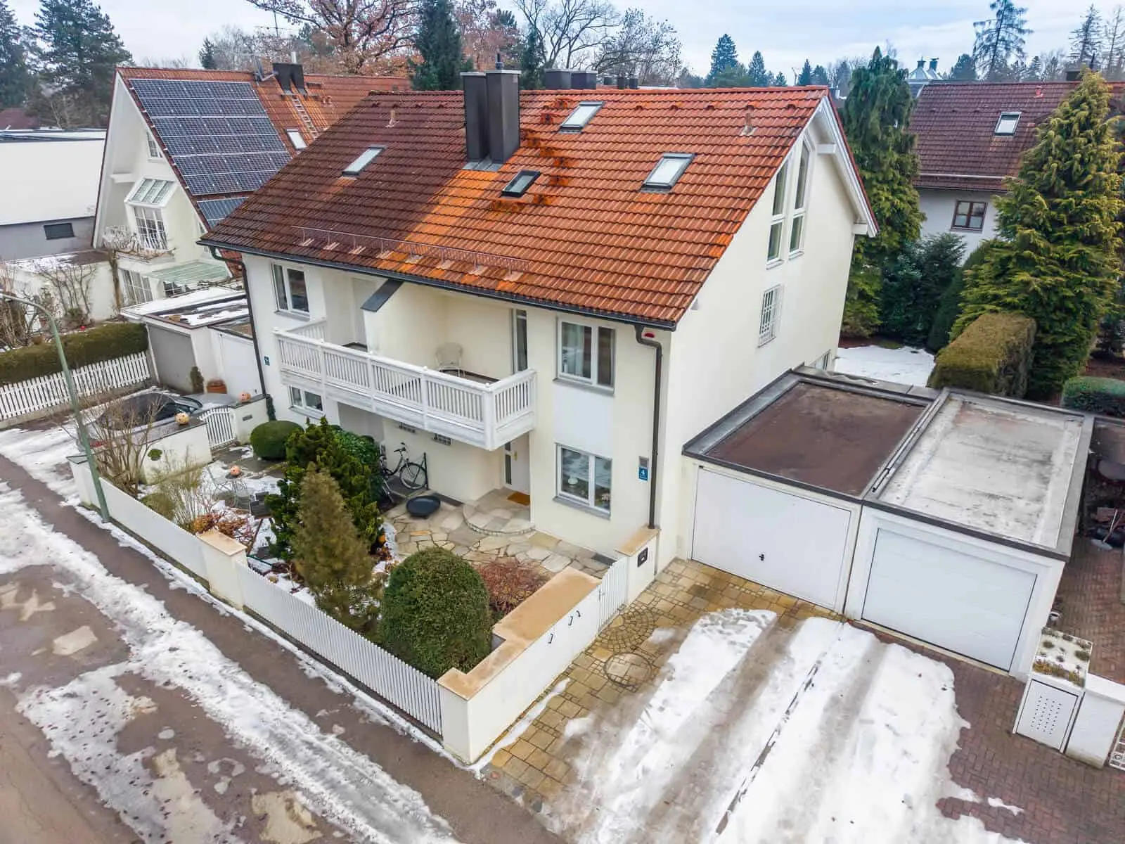 Ein zweistöckiges Haus mit rotem Ziegeldach, weißem Äußeren, angebautem Balkon, kleinem Vorgarten, Doppelgarage und Schneeflecken auf dem Boden.