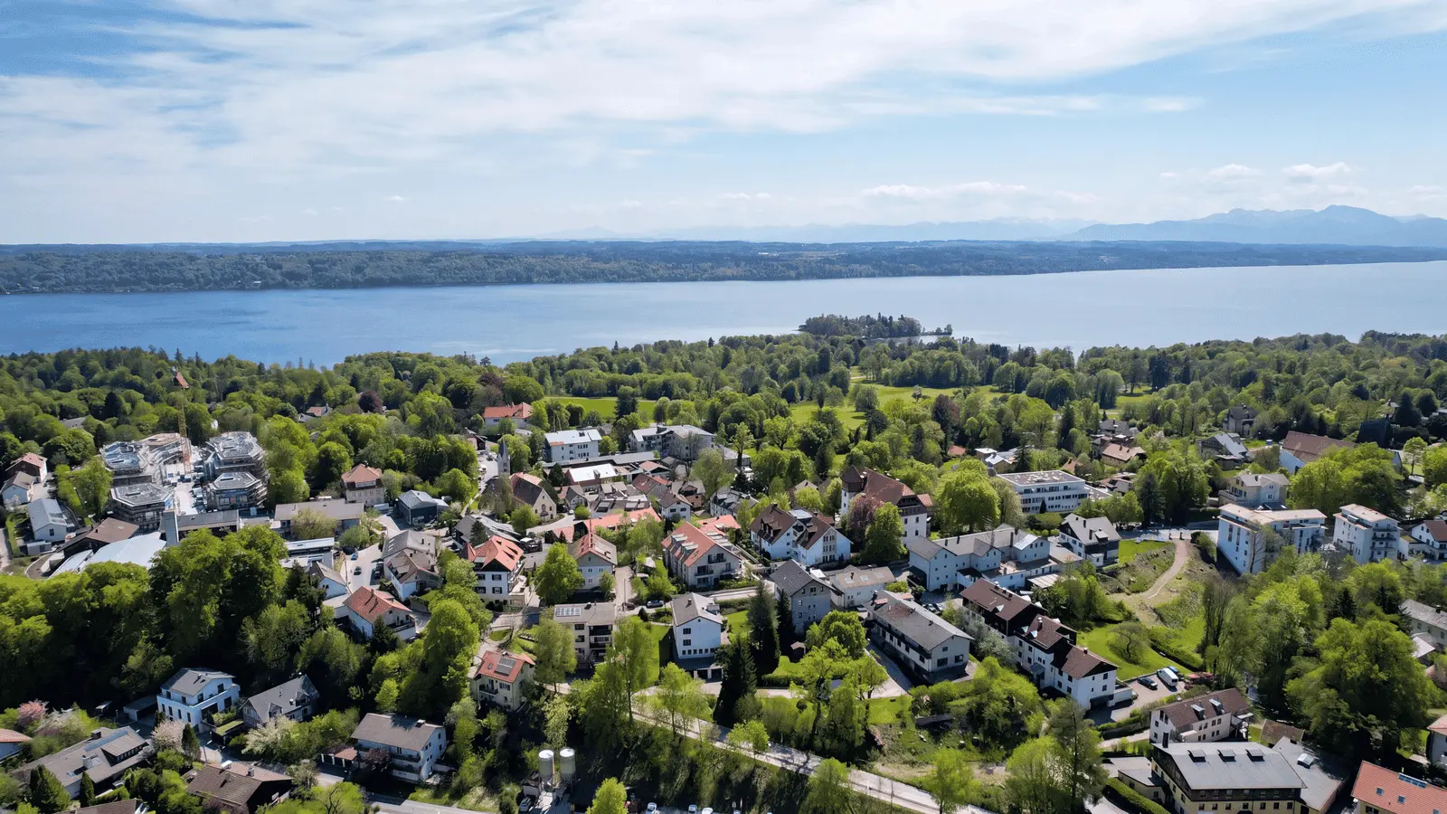 Luftaufnahme einer kleinen Stadt mit Häusern und Bäumen in der Nähe eines großen Sees, mit entfernten Hügeln und einem teilweise bewölkten Himmel im Hintergrund.