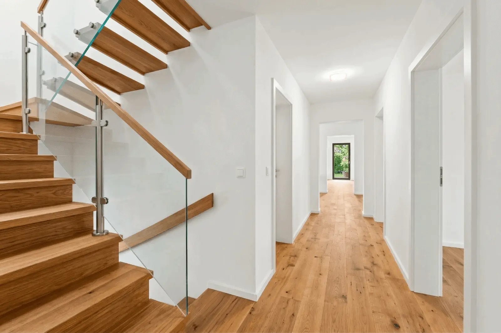 Moderner Flur mit Holzböden, weißen Wänden und einer schwebenden Holztreppe mit Glasgeländer auf der linken Seite; mehrere offene Türen und Tageslicht am Ende.