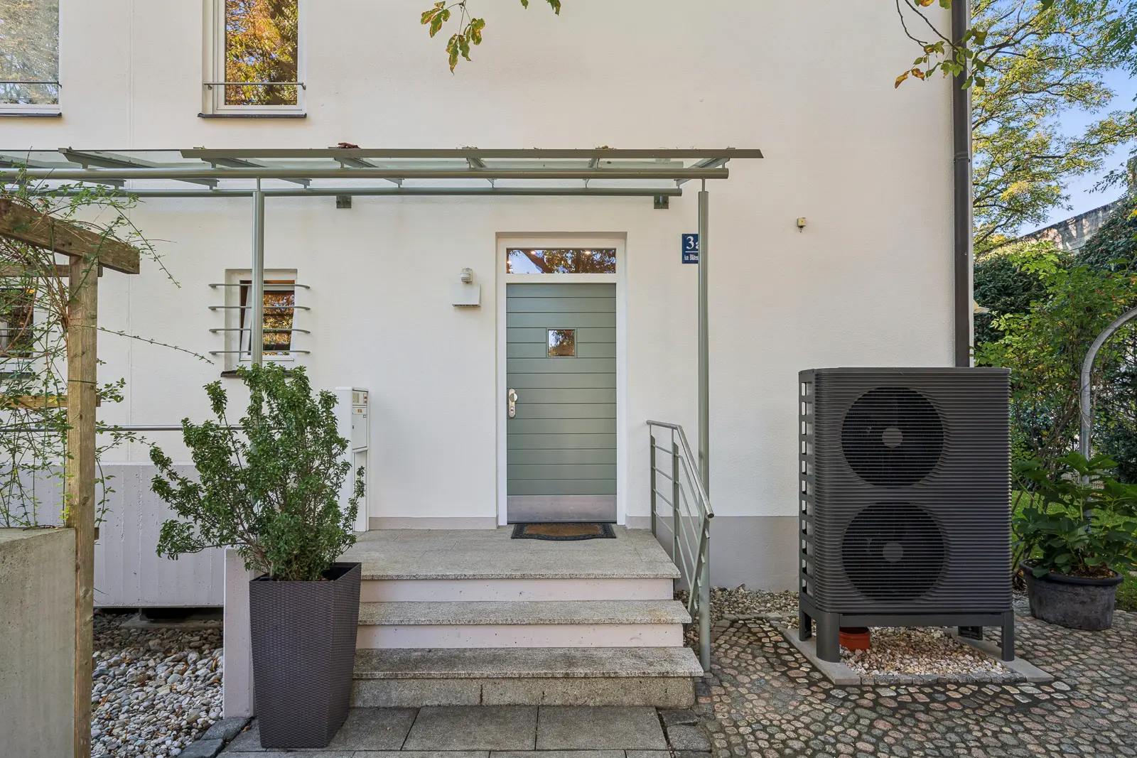 Der Haupteingang eines weißen Hauses mit einer grünen Tür, einem Glasvordach und einer großen schwarzen Wärmepumpeneinheit auf der rechten Seite.