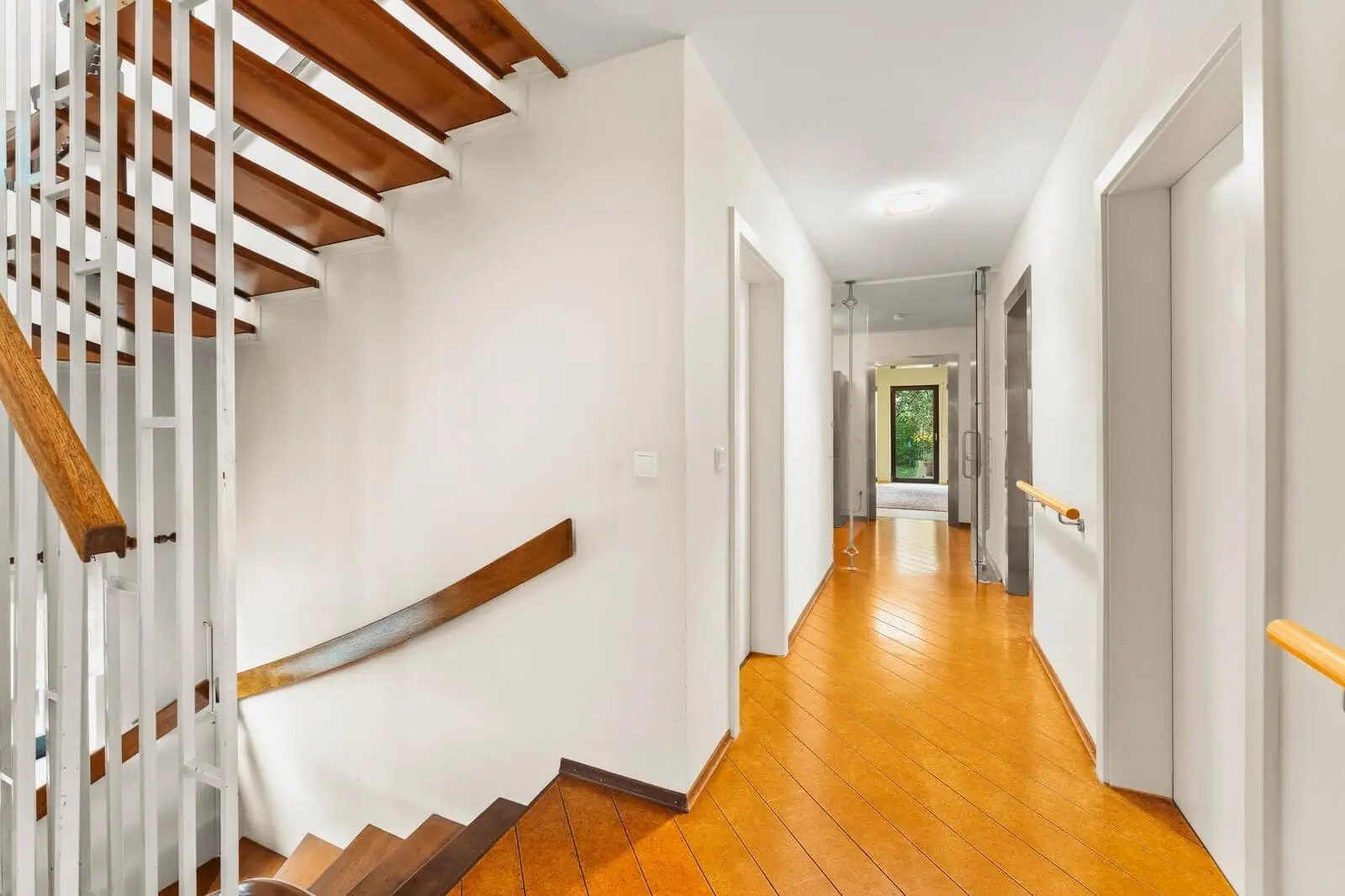 Ein heller Flur mit Holzböden, weißen Wänden, einer Holztreppe auf der linken Seite und Türen, die zu verschiedenen Räumen führen. Aus einem Zimmer am Ende des Flurs fällt natürliches Licht ein.