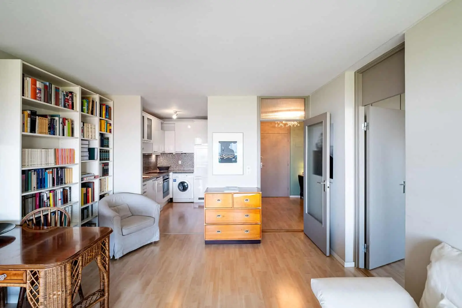 Helle Wohnungseinrichtung mit Bücherregalen, einem Holzschreibtisch, einem Sessel, einer Kommode und einem offenen Blick in eine moderne Küche und einen Flur.
