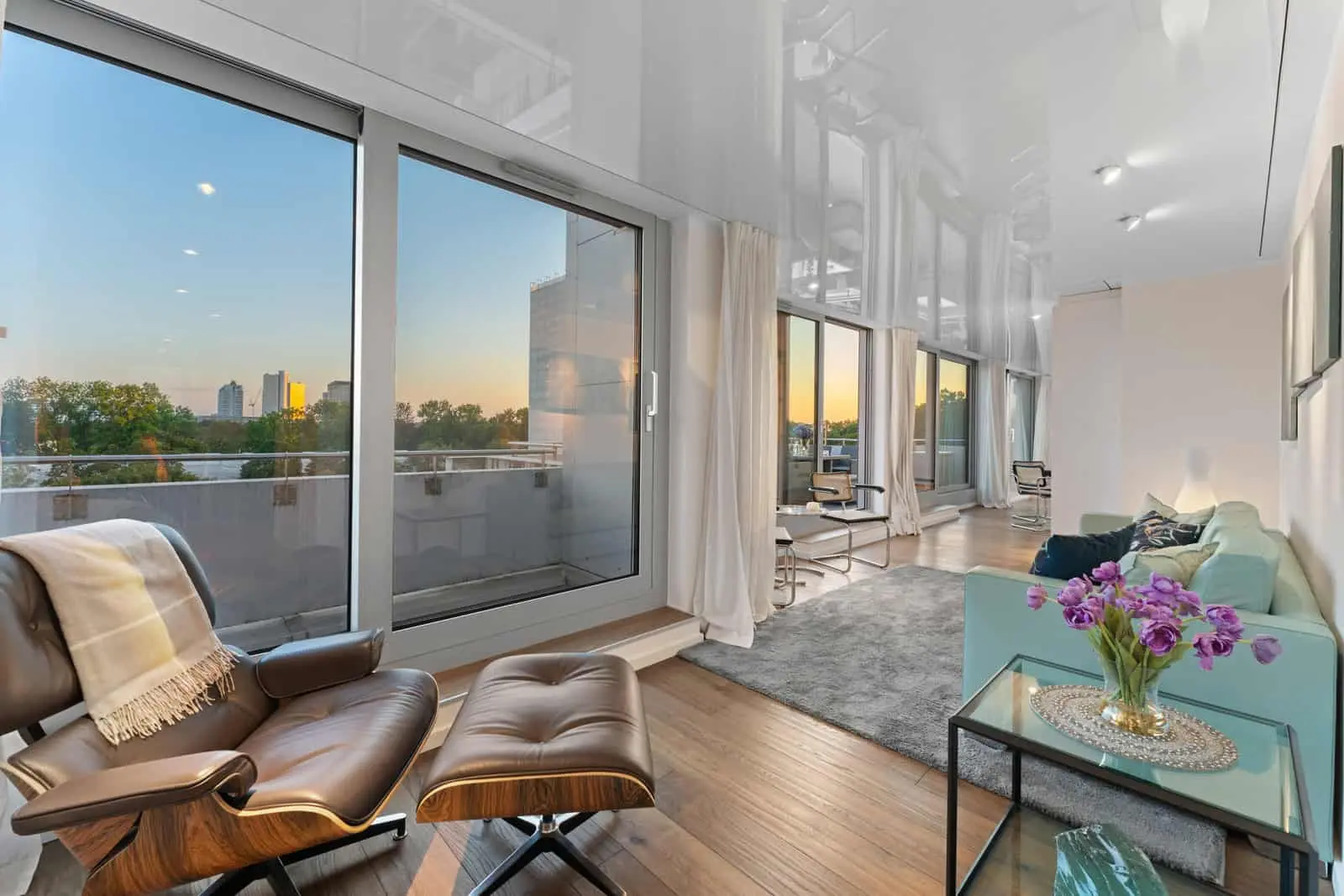Modernes Wohnzimmer mit großen Fenstern, Blick auf die Stadt, einem Loungesessel, einem hellblauen Sofa und einem kleinen Tisch mit lila Blumen, beleuchtet von natürlichem Sonnenuntergangslicht.
