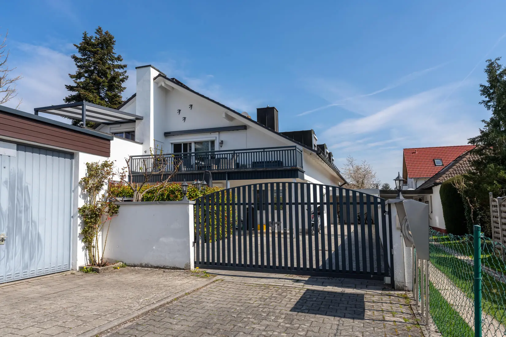Modernes zweistöckiges weißes Haus mit Balkon, schwarzem Tor, gepflasterter Auffahrt und einer Garage auf der linken Seite unter einem klaren blauen Himmel.