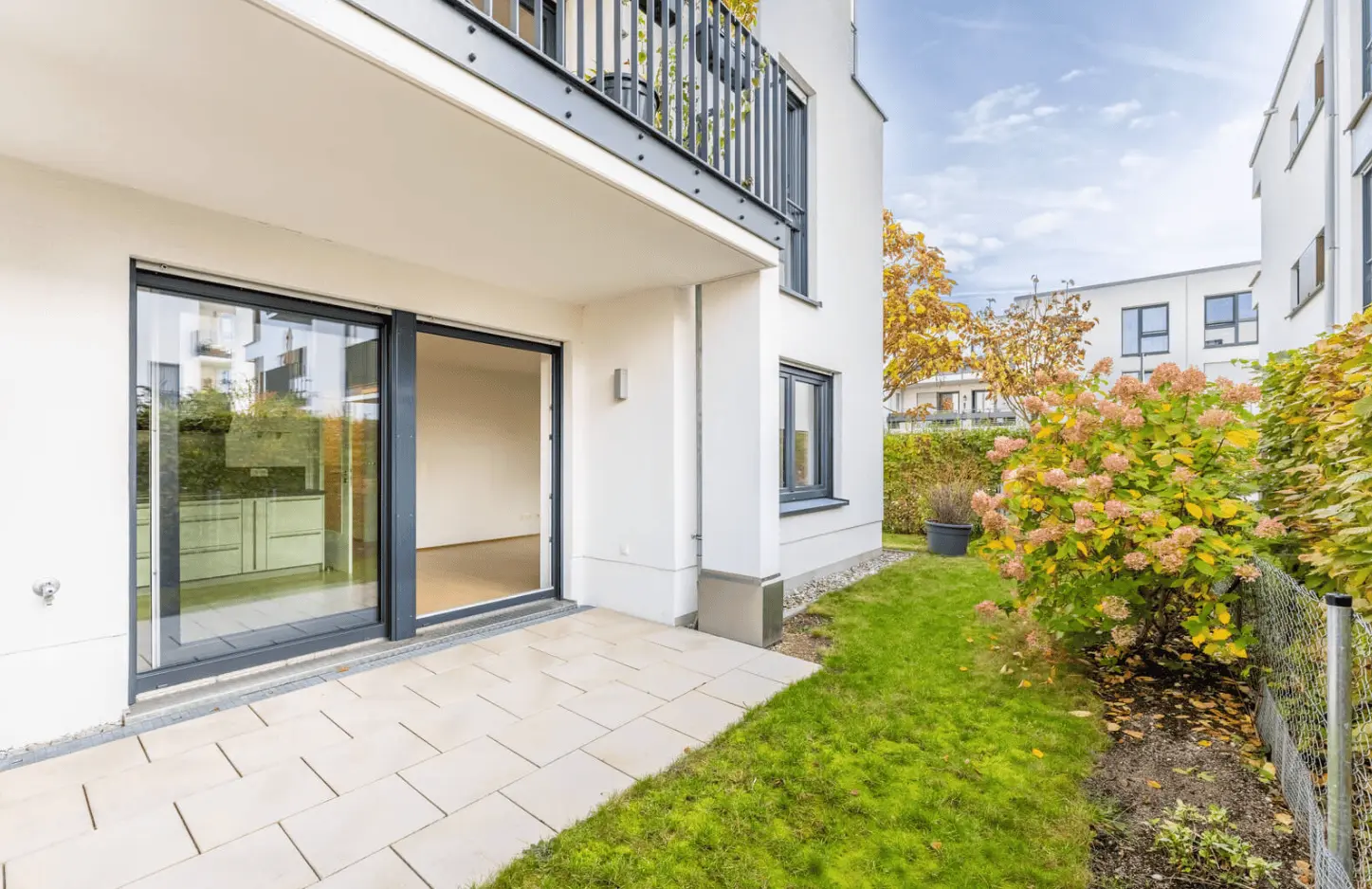 Wohnung im Erdgeschoss mit Glasschiebetüren, die auf eine geflieste Terrasse und einen kleinen grasbewachsenen Garten führen, umgeben von Büschen und modernen Wohngebäuden.