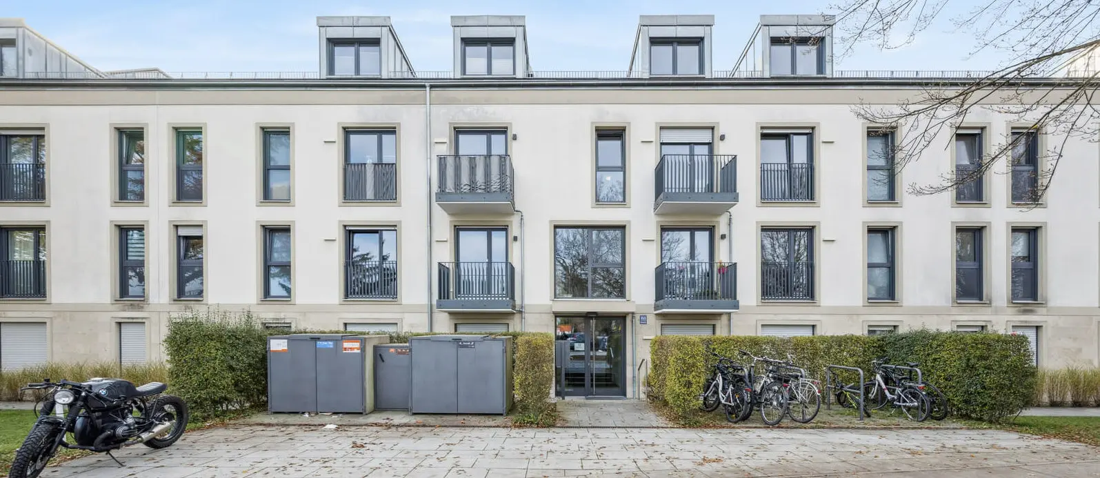 Ein modernes dreistöckiges Wohnhaus mit Balkonen, Fahrradständern, Mülleimern und einem geparkten Motorrad davor, umgeben von Herbstlaub.