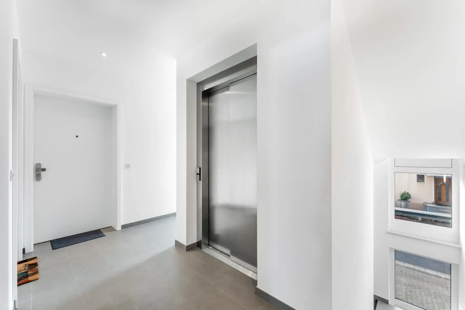 Ein moderner Wohnhausflur mit einer geschlossenen Aufzugstür, einer weißen Eingangstür, hellgrauen Bodenfliesen und einem Fenster an der Treppe.