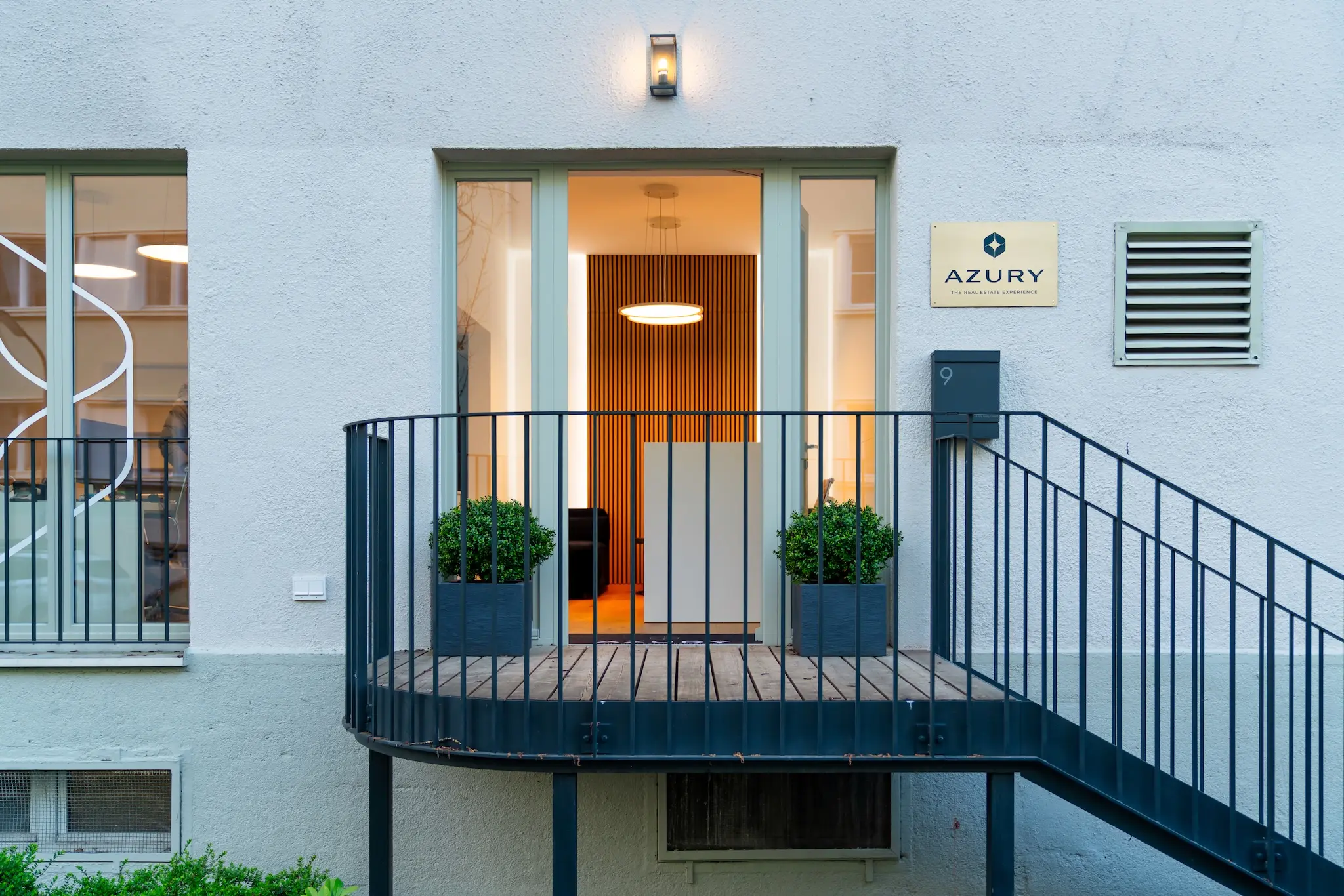 Kleiner moderner Gebäudeeingang mit einer schwarzen Metalltreppe, Glastüren, zwei Topfpflanzen und einem Schild mit der Aufschrift „AZURY“ an der Wand.