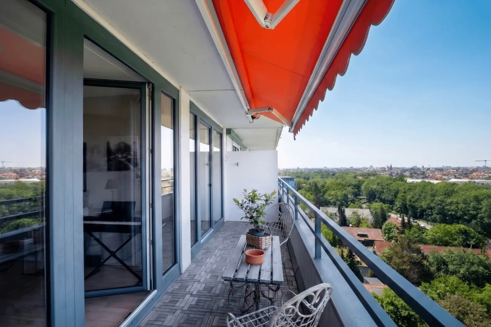 Ein moderner Balkon mit Glastüren, Holzboden, Metallstühlen, einem kleinen Tisch mit einer Topfpflanze und einer orangefarbenen Markise mit Blick auf eine grüne Landschaft und das Stadtbild.