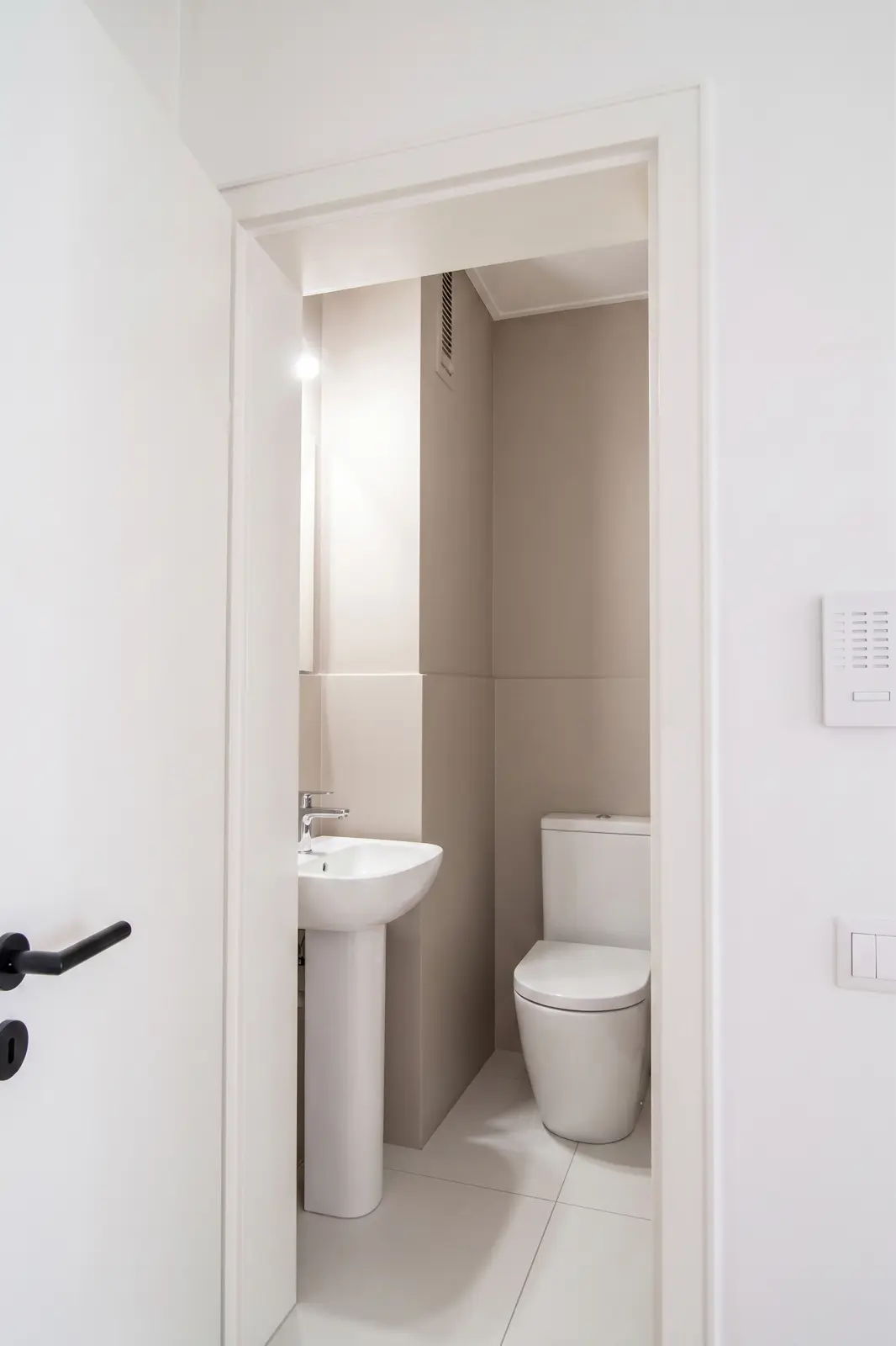 Kleines, modernes Badezimmer mit einer weißen Toilette und einem Standwaschbecken, beigen Wänden und einer teilweise geöffneten weißen Tür.