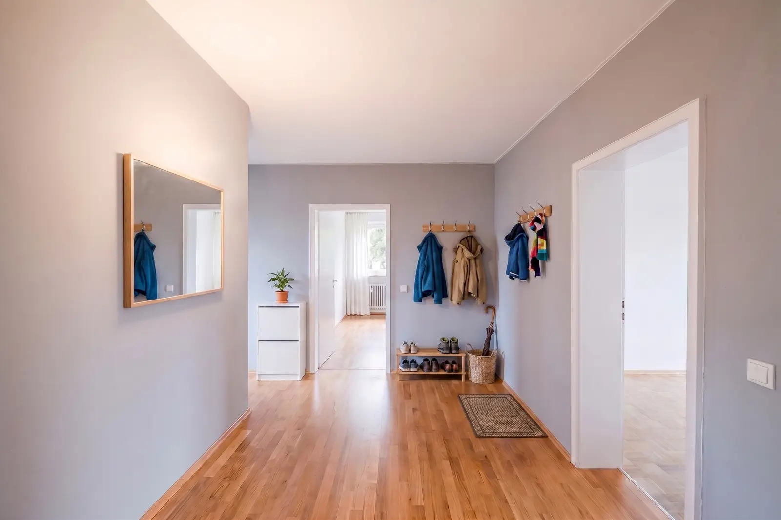 Ein aufgeräumter Flur mit Holzboden, Garderobenstangen mit Jacken, einem Schuhregal, einem Spiegel an der Wand, einem kleinen Schrank mit einer Pflanze und Türen, die zu anderen Räumen führen.