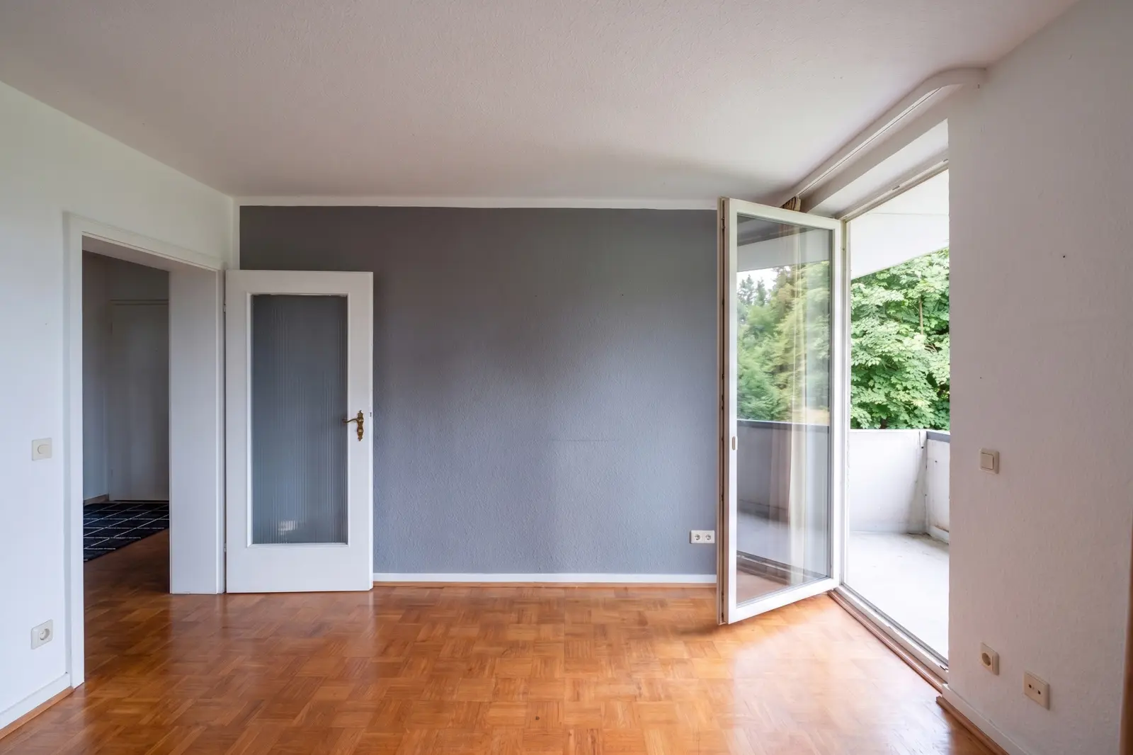 Leerer Raum mit Parkettboden, hellgrauer Akzentwand, offener Tür zum Flur und offener Glastür, die auf einen Balkon mit Blick ins Grüne führt.